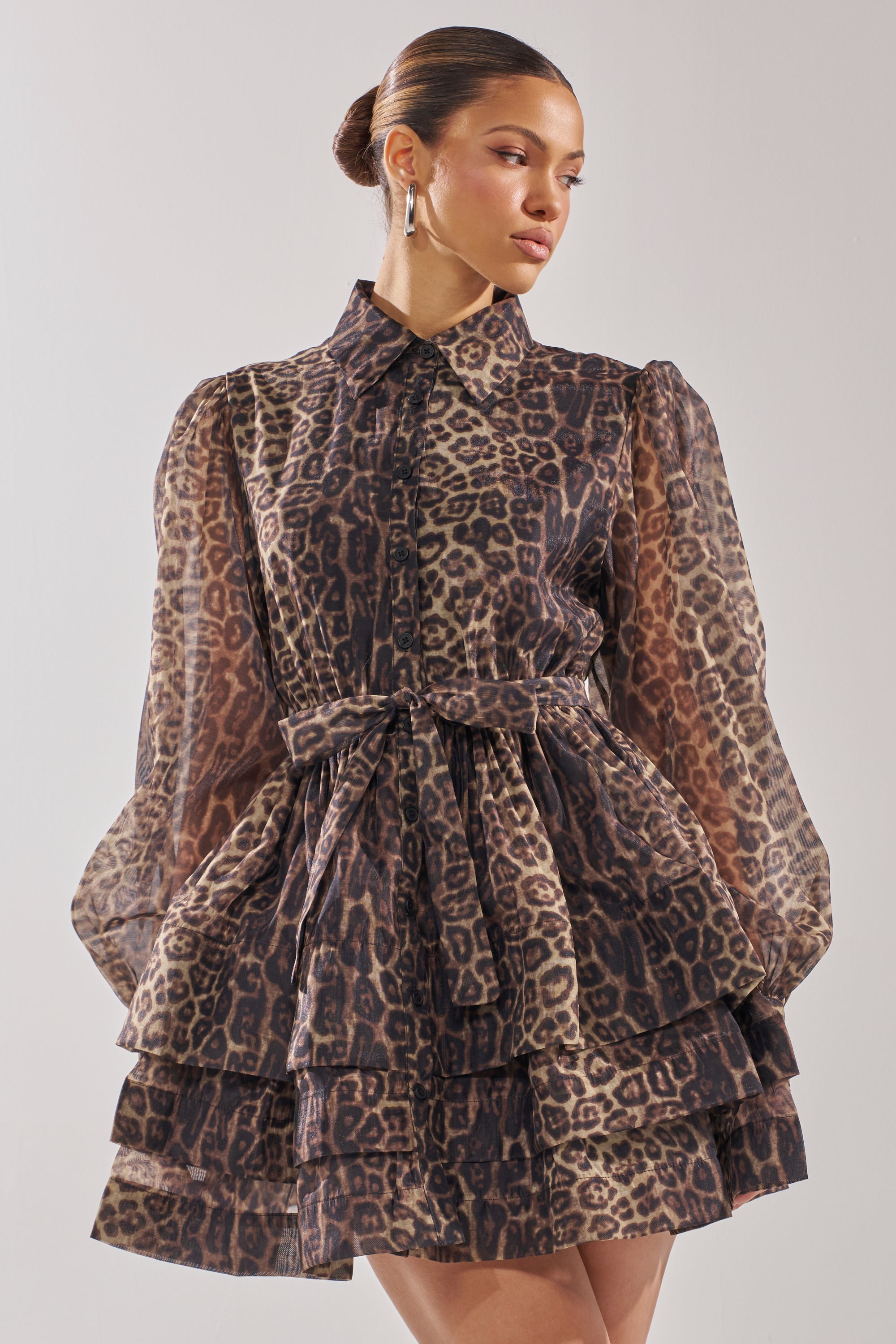 A woman wearing the CHICEST OF CHIC LEOPARD PRINT MINI DRESS, featuring sheer long sleeves, a collared neckline, a tied waist, and tiered skater silhouette, stands against a plain light background.