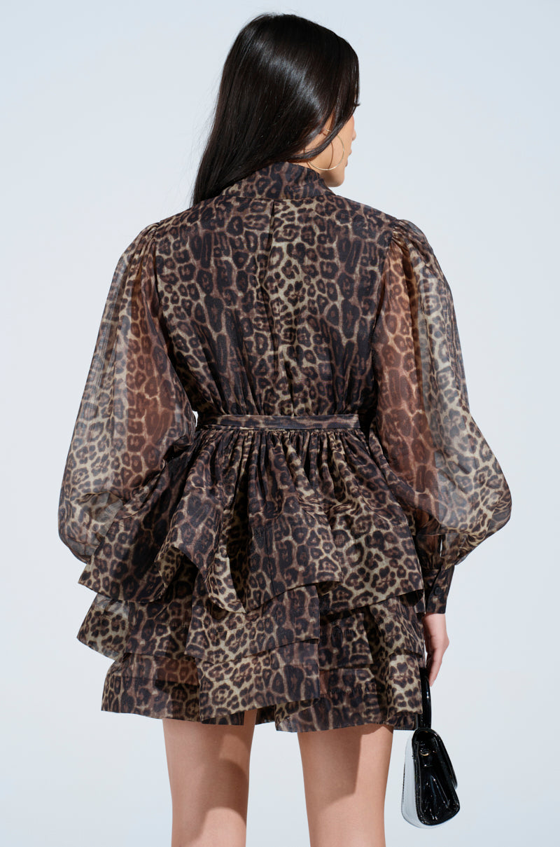 Against a simple light gray background, a person with long dark hair is seen from the back wearing the CHICEST OF CHIC LEOPARD PRINT MINI DRESS with bishop sleeves, holding a small black handbag.