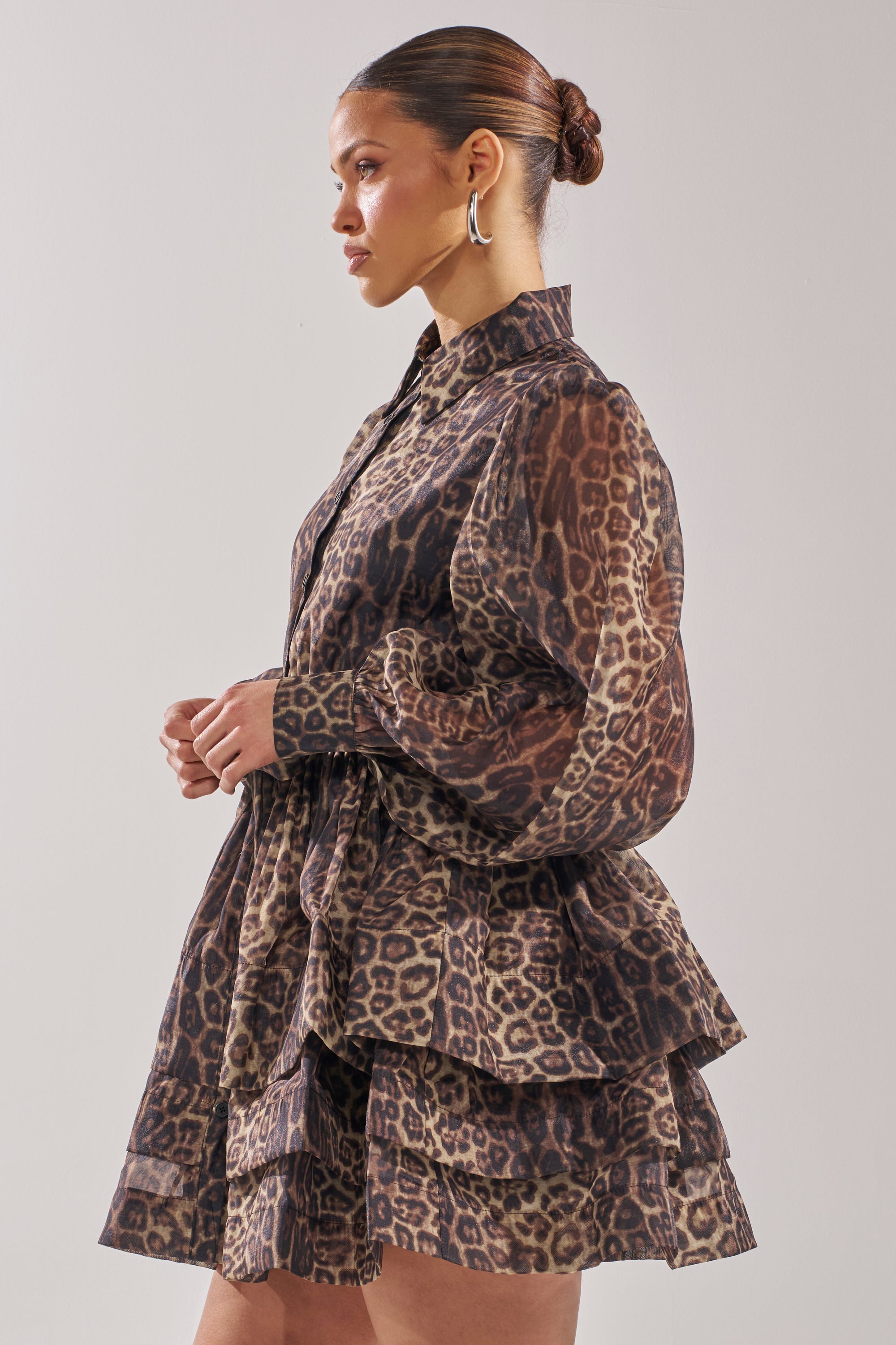 A woman with her hair in a bun stands in profile against a plain light background, wearing the Chicest of Chic Leopard Print Mini Dress with long sleeves and a skater silhouette.
