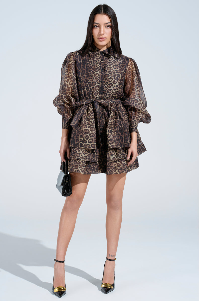 A woman wearing the Chicest of Chic Leopard Print Mini Dress with bishop sleeves and a bow tie waist, accessorized with a small black handbag and black and gold heels, stands against a light background. Her long, straight hair flows elegantly.
