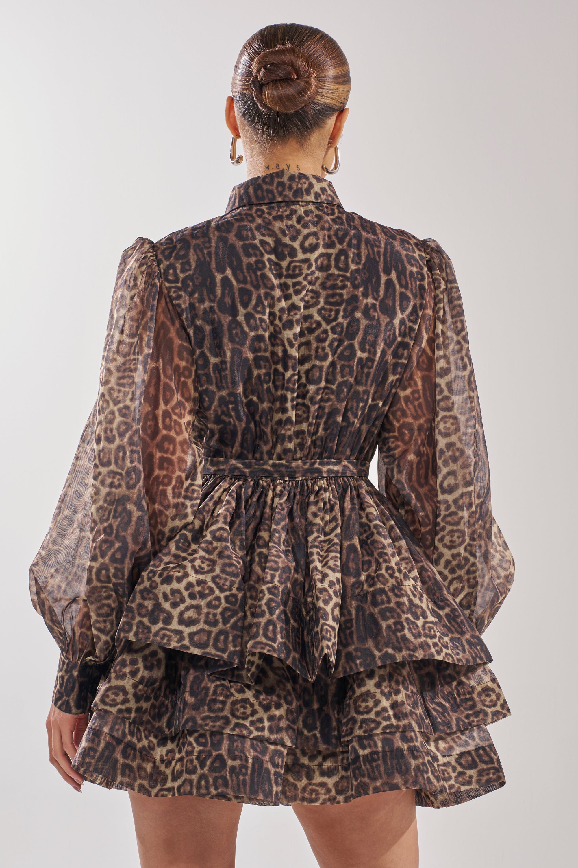 A woman with her hair in a sleek bun is seen from the back wearing the CHICEST OF CHIC LEOPARD PRINT MINI DRESS, featuring tiered ruffles, sheer long sleeves, and a skater mini dress silhouette.
