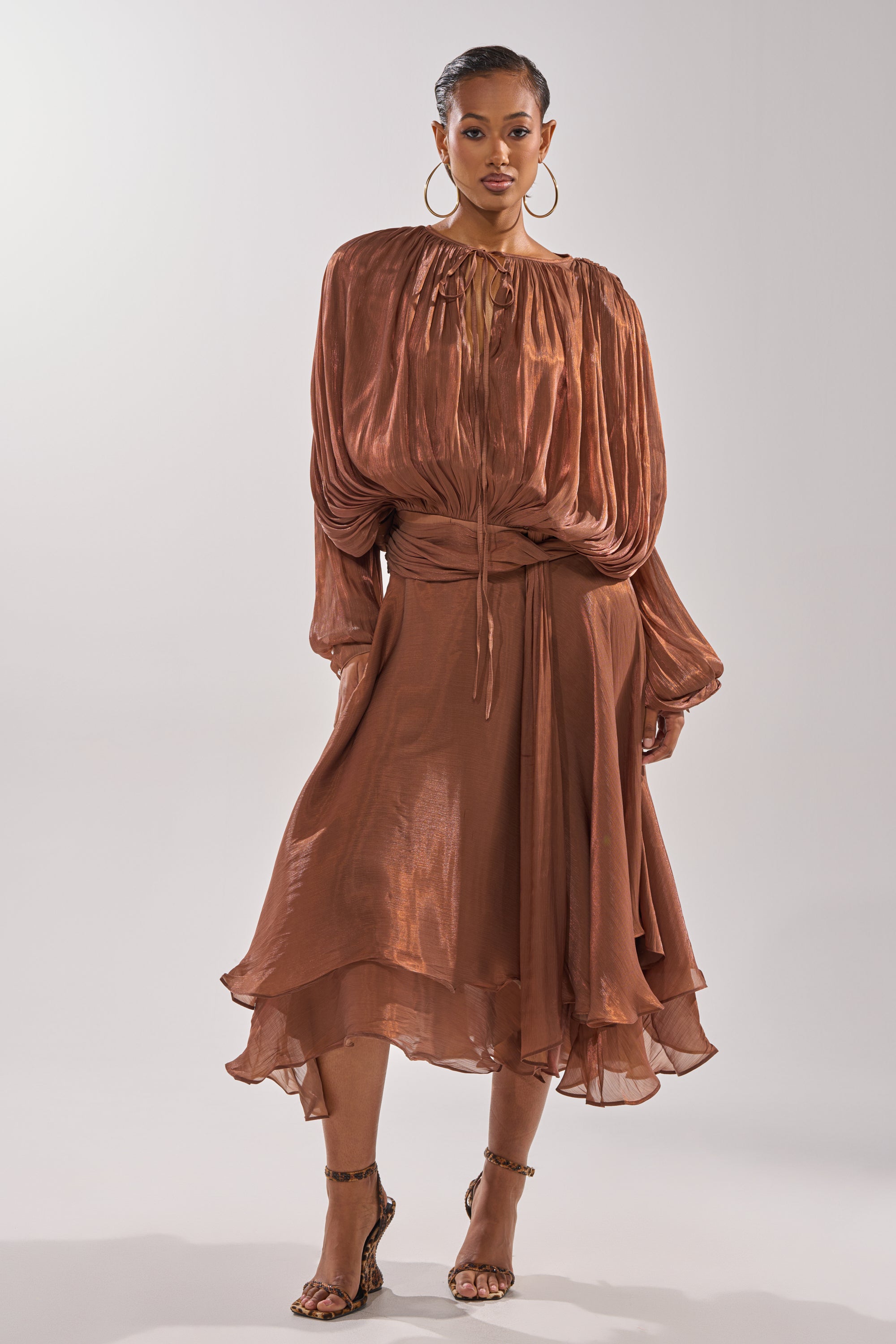 A woman exudes confidence in the GINA MAXI DRESS, a shiny copper dress with layered, flowy fabric and a flattering waist tie. She elevates the look with hoop earrings and strappy heels for a chic, elegant style. 1