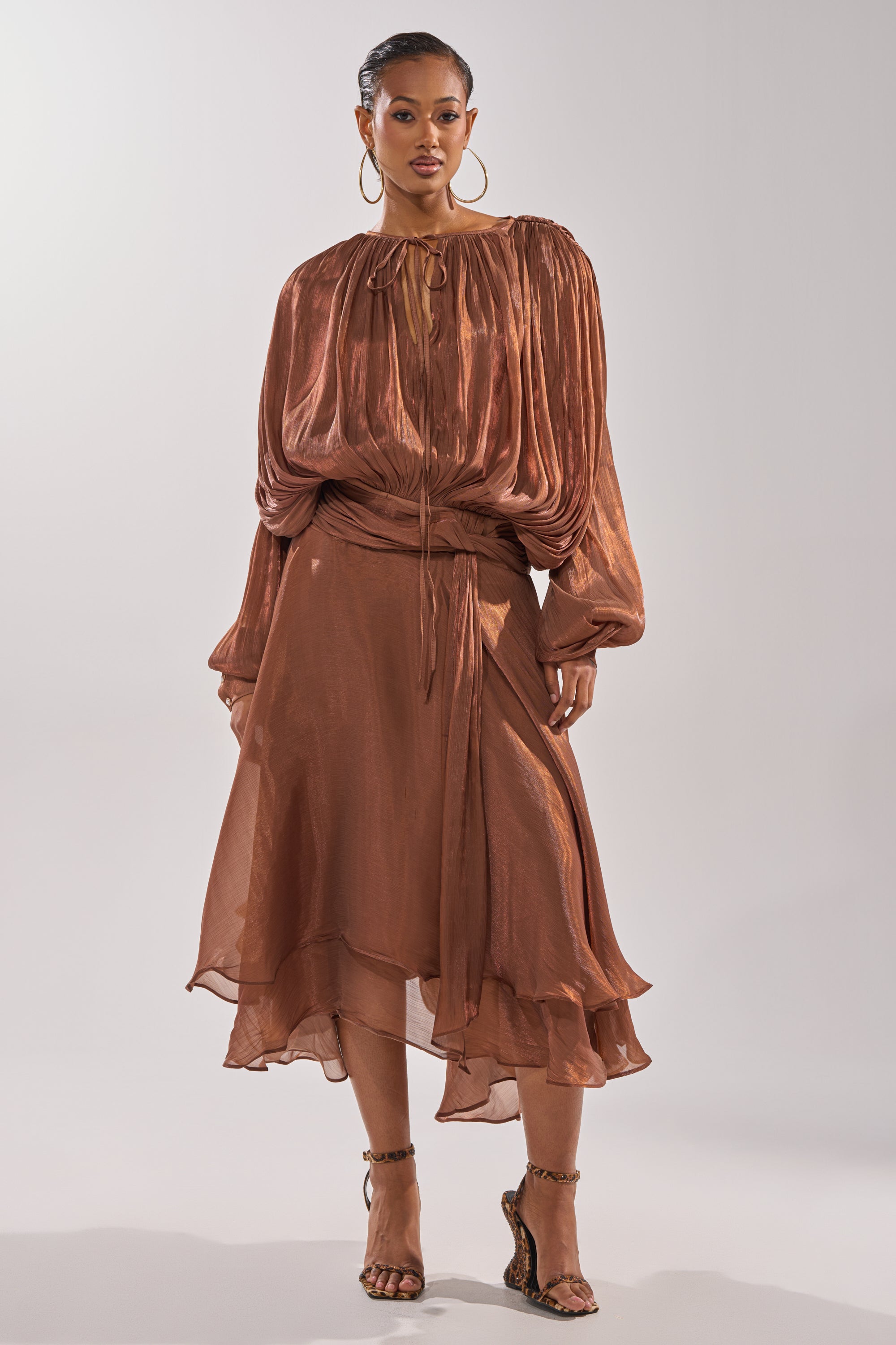 A woman elegantly poses against a plain light background, showcasing the refined charm of the GINA MAXI DRESS in copper with pleated details, capturing sophisticated style in one stunning piece. 2