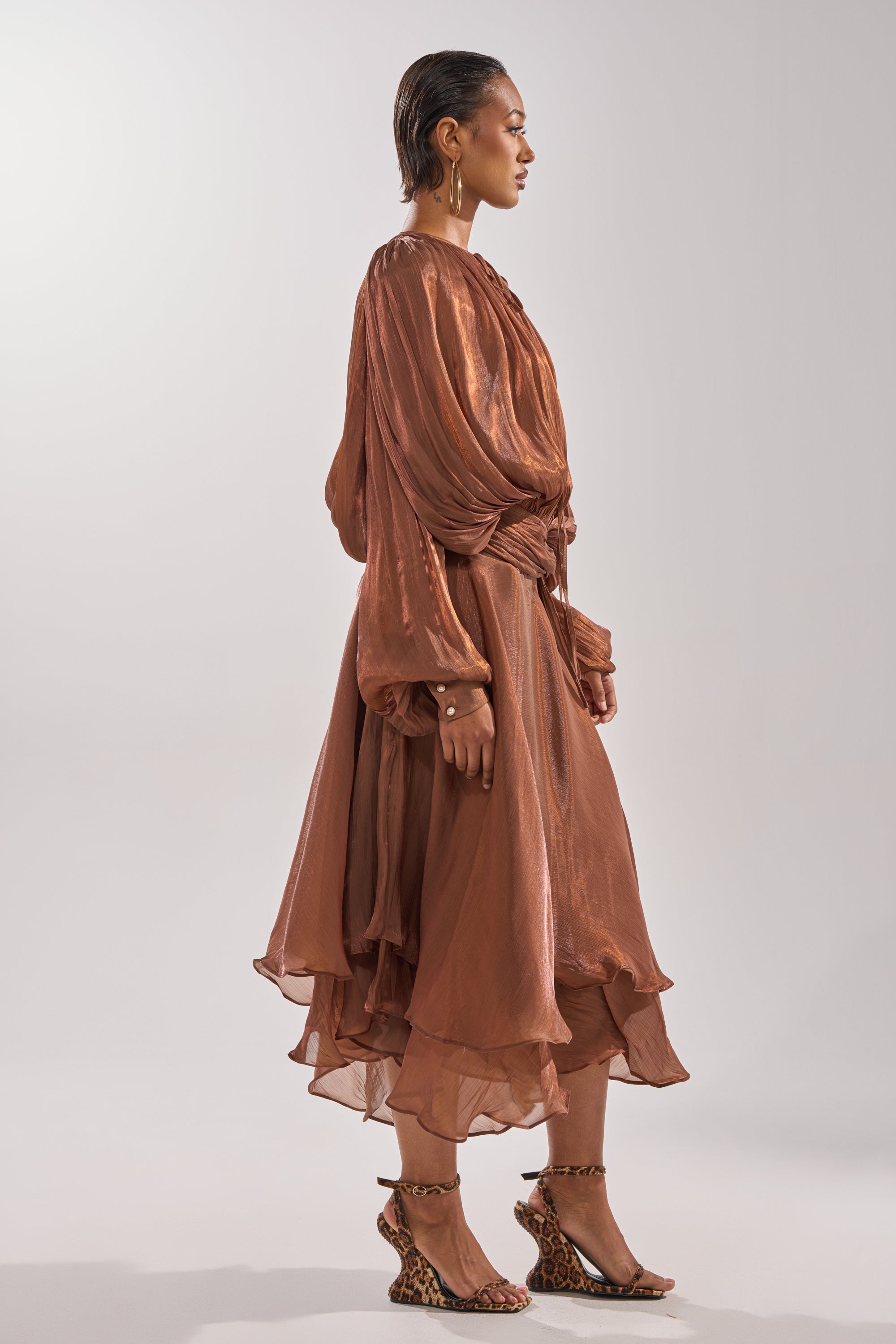 A person stands sideways in the GINA MAXI DRESS, a flowy copper-brown, long-sleeve chiffon dress with ruffled layers and knee-length hem, paired with leopard-print heeled sandals against a plain light background. 3