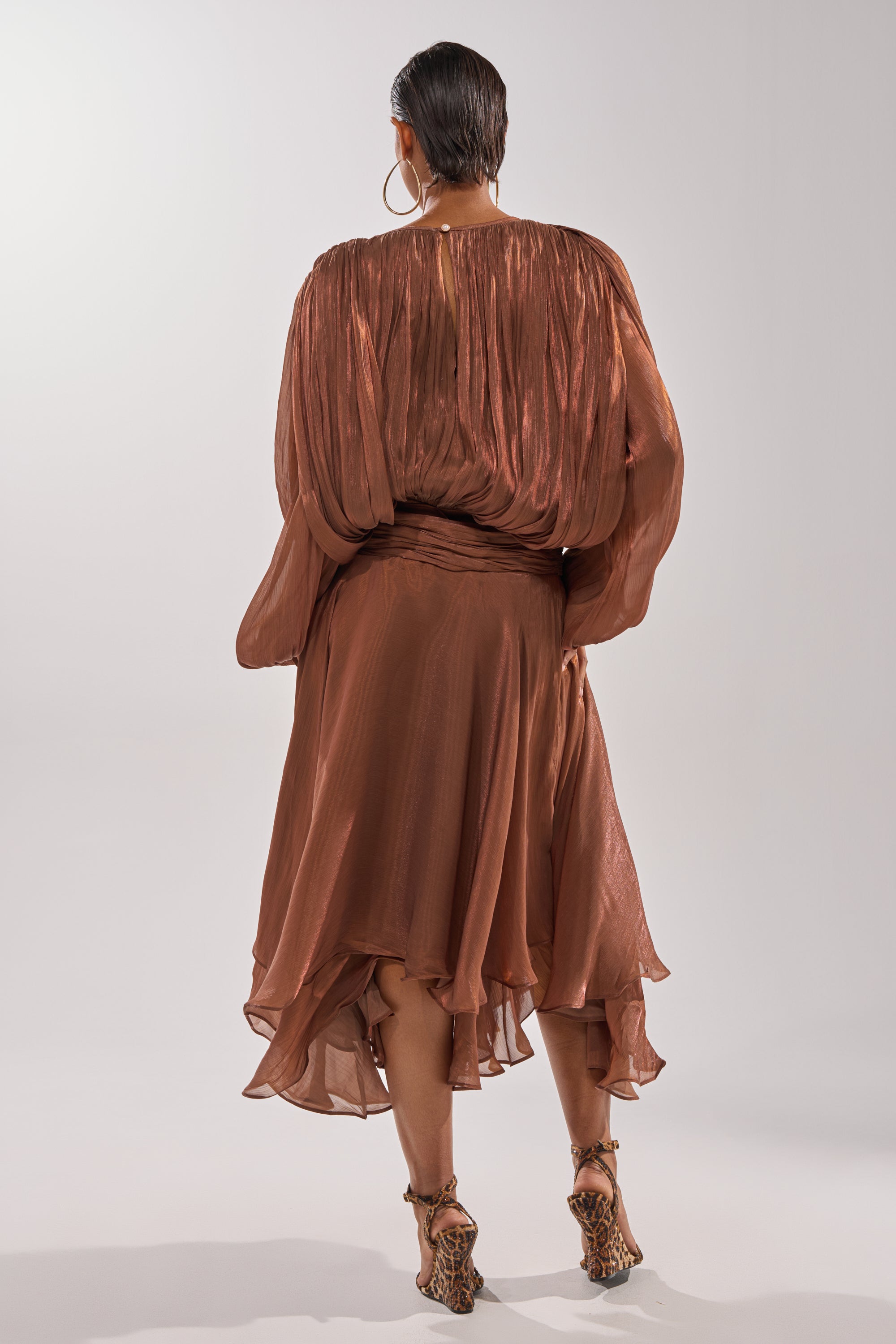 A person stands with their back to the camera, wearing the shiny copper GINA MAXI DRESS with ruffled edges and strappy sandals, radiating effortless elegance. 4