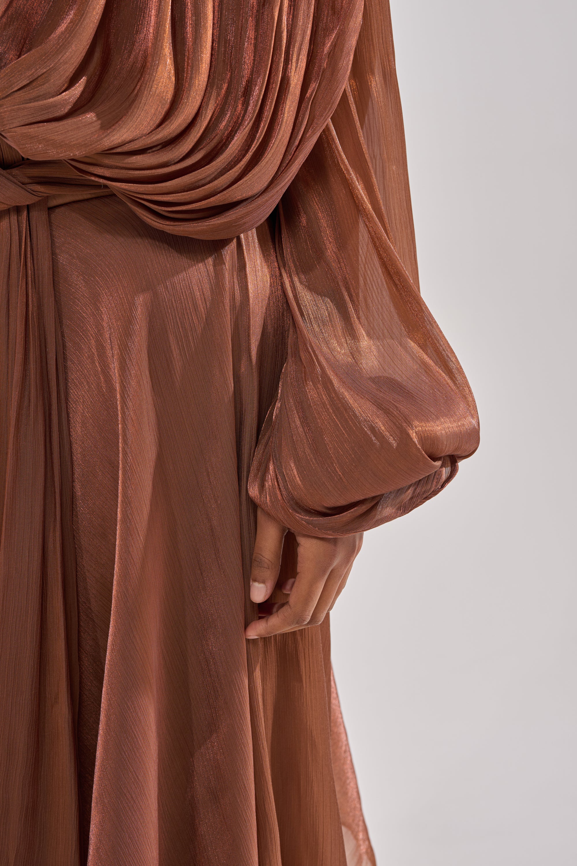 A close-up of a person wearing the GINA MAXI DRESS, a flowy sheer brown chiffon maxi dress with draped fabric and long, billowy sleeves. One hand rests by their side against a neutral background. 5
