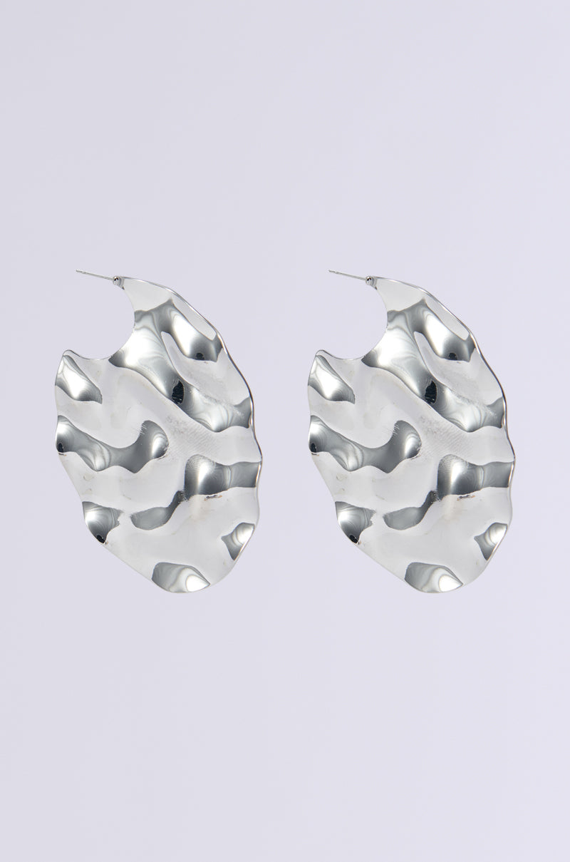 Introducing the CHILL WITH ME EARRING: a pair of statement jewelry earrings in metallic faux silver, featuring an oversized abstract wave design. These irregularly shaped earrings boast a hammered, textured surface, adding a distinctive artistic flair against a plain, light background.
