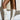 A person stands against a white brick wall, wearing a light beige coat and AZALEA WANG CECILE BONE SANDAL STILETTOS. Their legs are bare, with the coat draping behind them and casting shadows on the wall and floor.