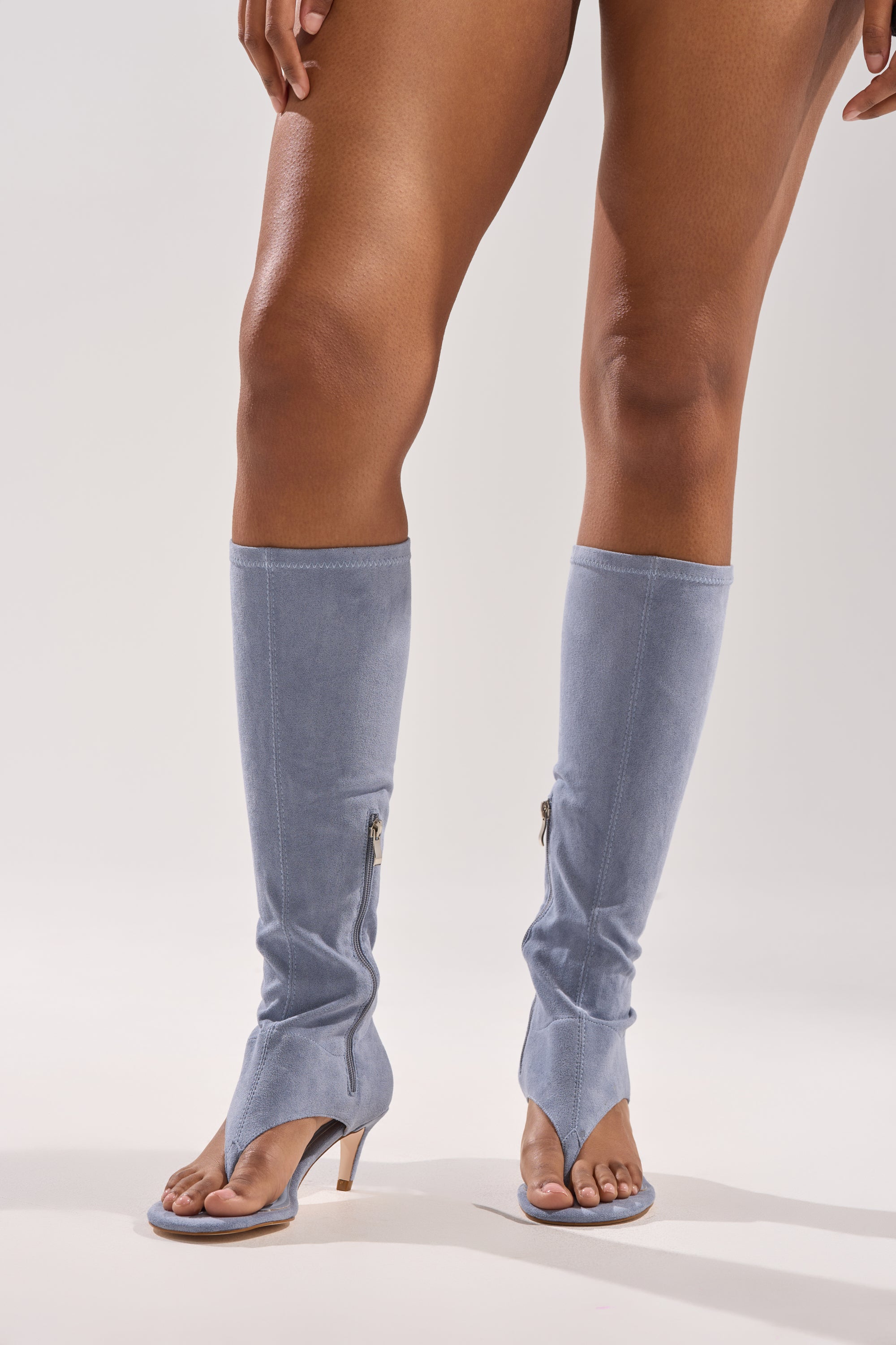 A person stands on a plain light background wearing AZALEA WANG CECILE LIGHT BLUE FAUX SUEDE SANDALS—knee-high, open-toe heeled boots with side zippers, a split-toe design, and stylish mid-height heels. 1
