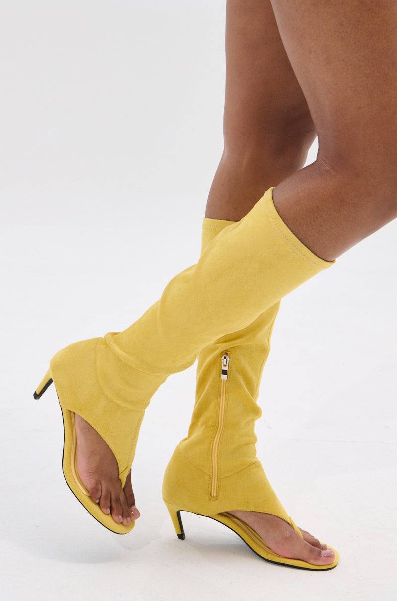 A person wearing the AZALEA WANG CECILE MUSTARD SANDAL STILETTO, posing with one leg crossed in front of the other against a white background.