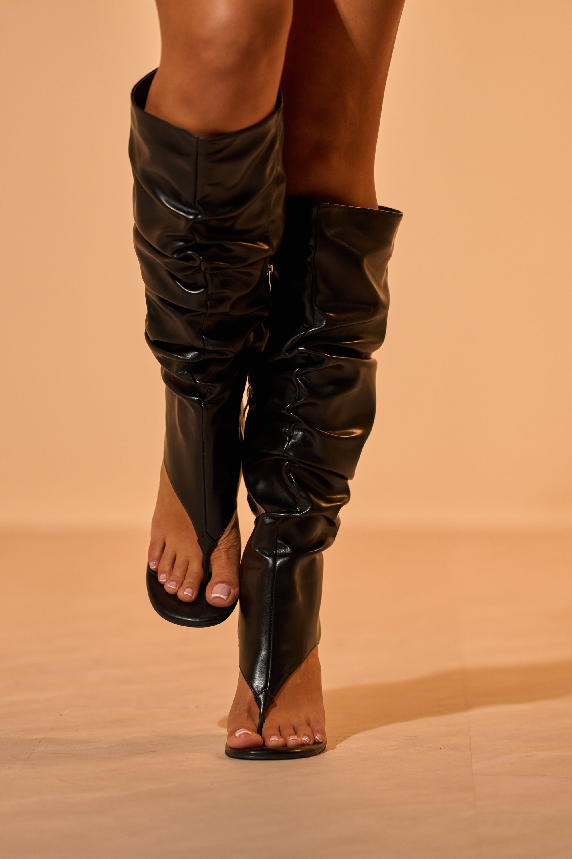 A person wearing AZALEA WANG CECILL BLACK RUCHED UPPER GLADIATOR SANDALS with tall, black, ruched faux leather uppers and open toes stands on a light beige floor against a neutral background. 1