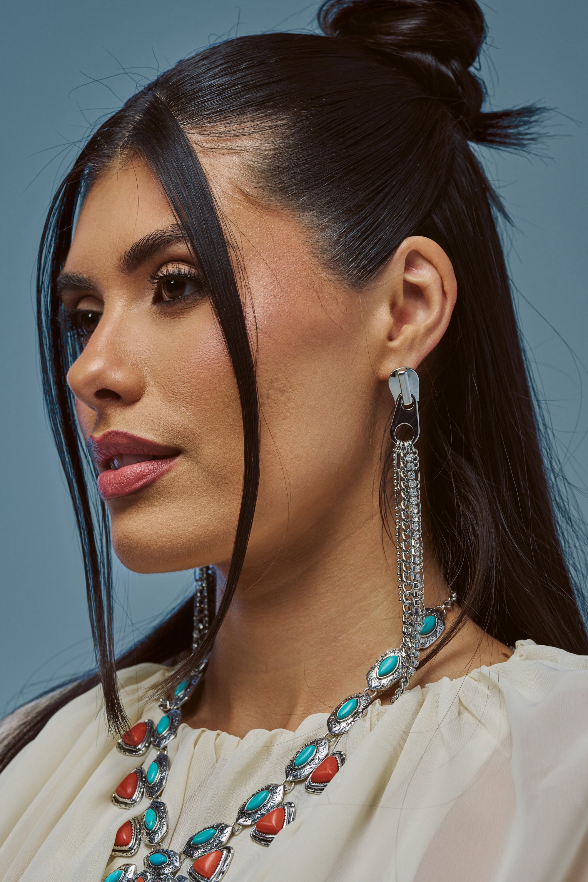 Woman with long dark hair styled half-up, wearing a cream top and the ZIP IT STATEMENT EARRING, poses against a plain blue background.