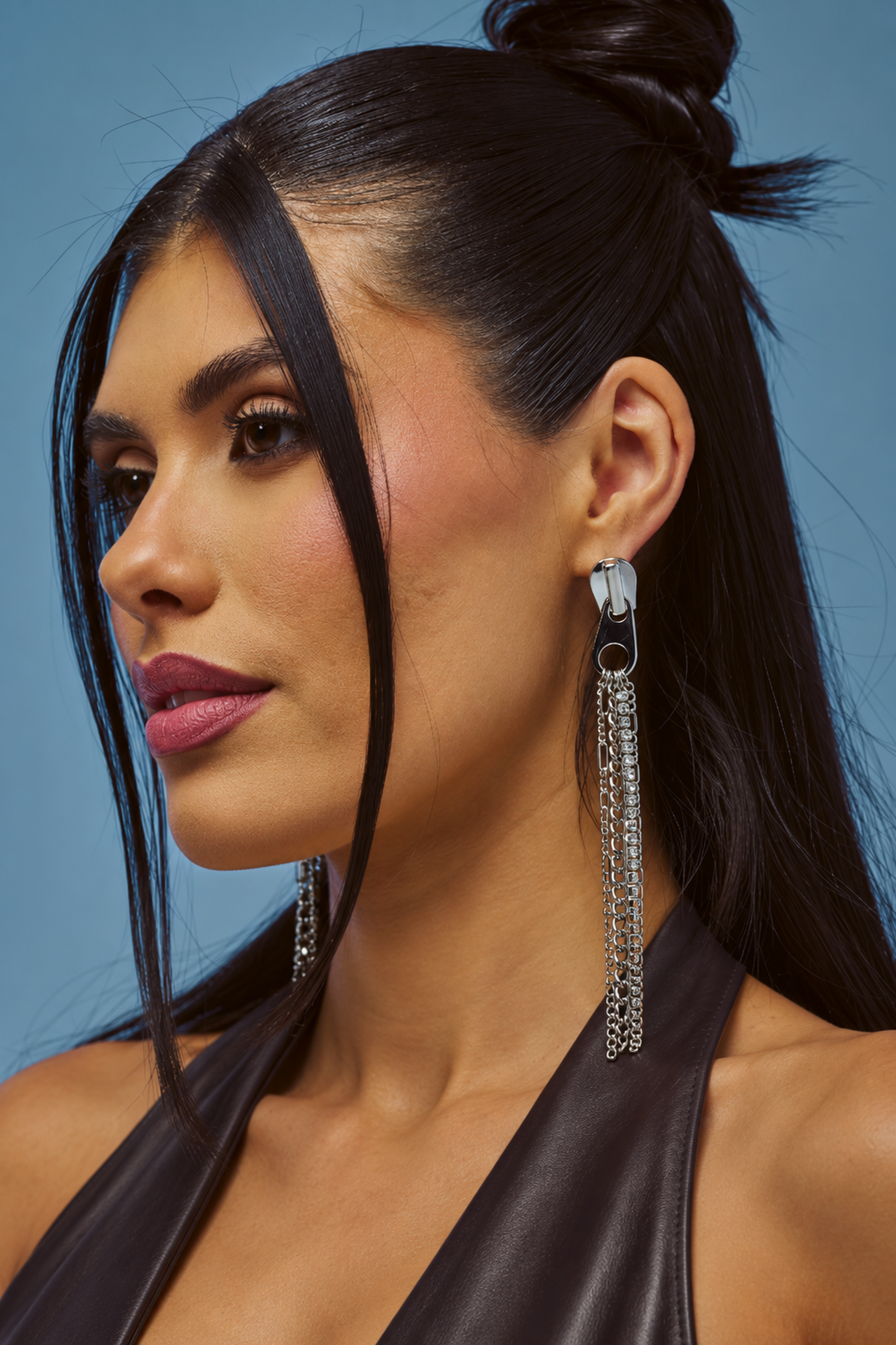 Woman with long dark hair styled half-up wears the ZIP IT STATEMENT EARRING—bold, silver chain earrings—and a black sleeveless top, posing against a blue background.