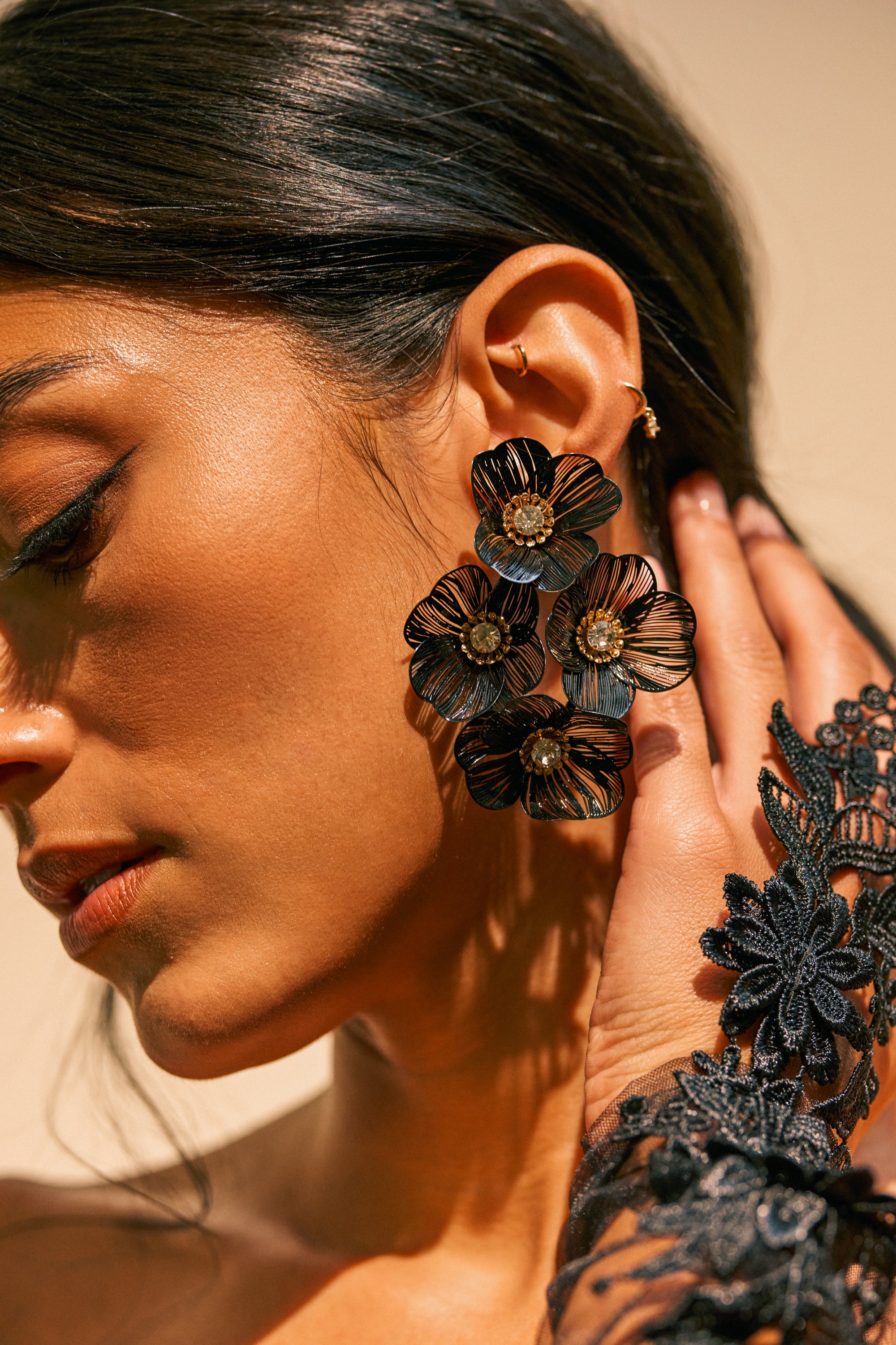 A woman wearing the BLACK BLOSSOM DRAMA EARRINGS—striking, oversized black floral clusters—pairs them with a black lace glove as she touches her ear; her dark hair is pulled back.