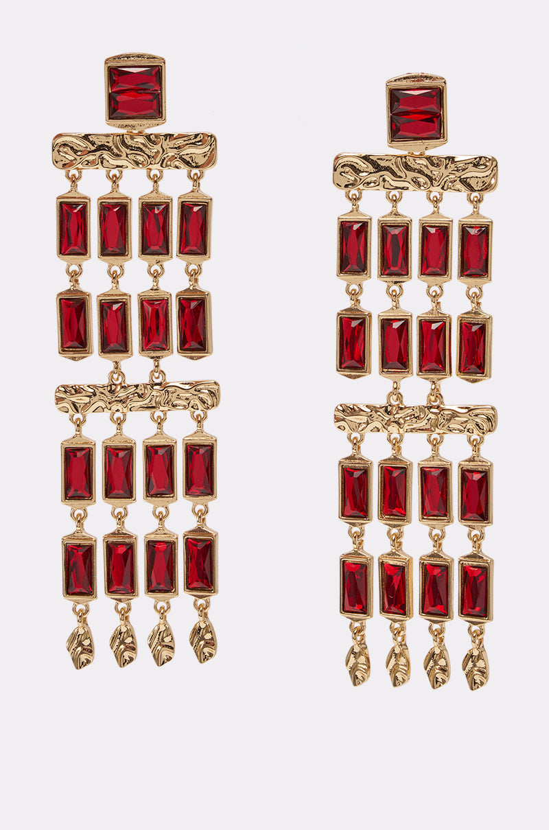 The BAGUETTE CHANDELIER EARRINGS IN RED feature multiple rectangular red gemstones and small gold leaf charms dangling at the bottom, making these gold statement earrings a striking accessory, shown here on a white background.