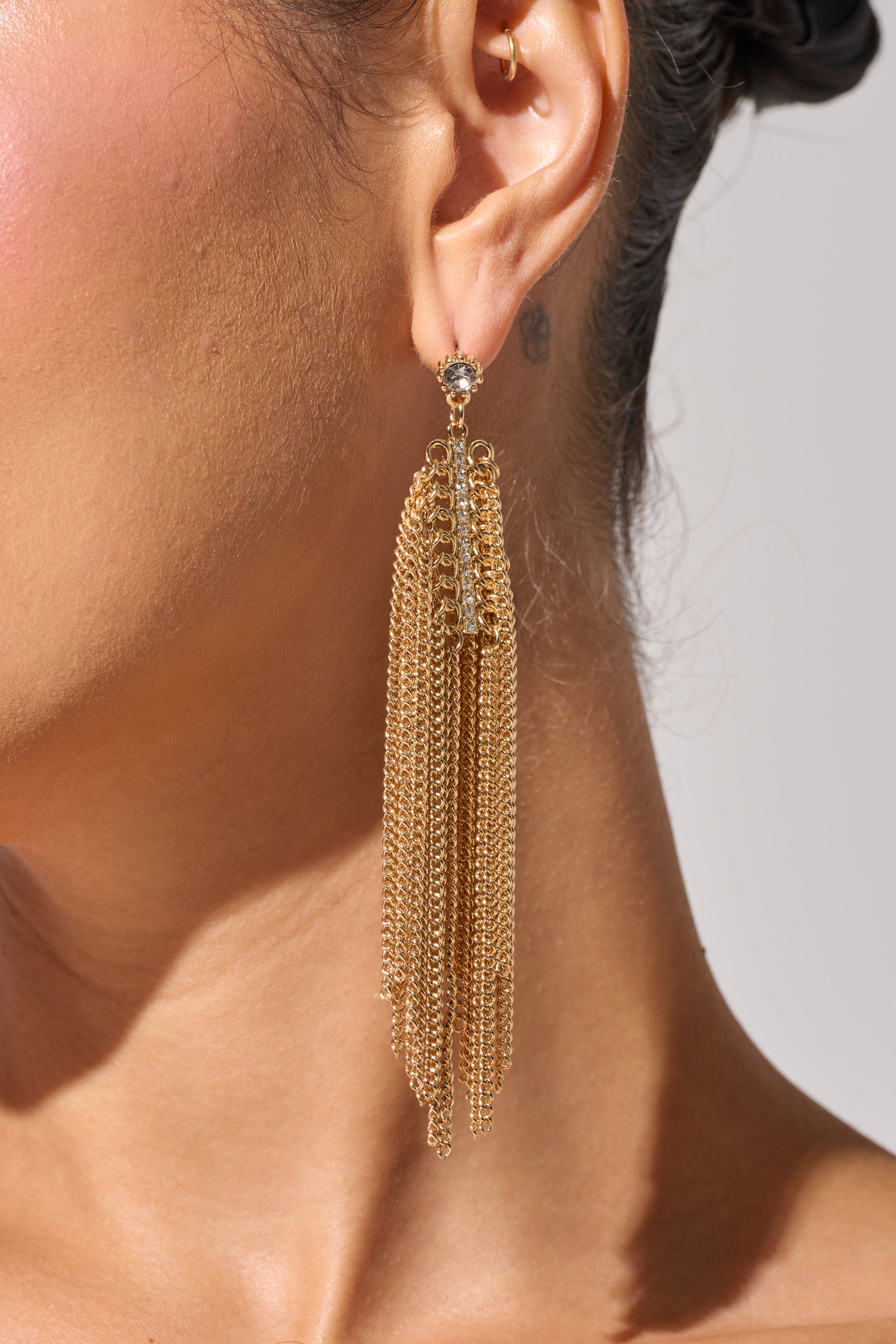 Close-up of a woman's ear wearing the SHAKE IT EARRING—long, gold-tone earrings featuring multi-strand cascading chain fringe. A neutral background with part of her neck and shoulder is visible. 1