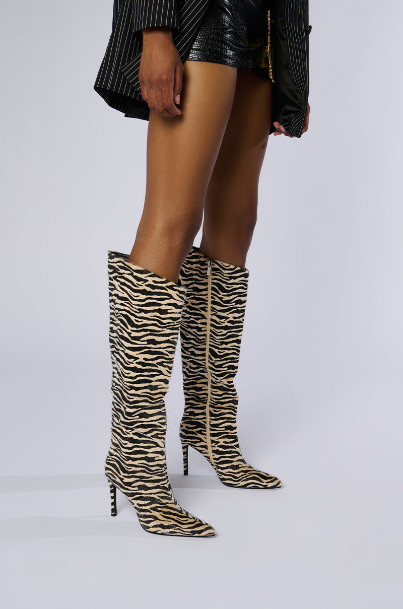 A person is wearing the AZALEA WANG MULTI PONY HAIR ZEBRA BOOT, which are knee-high, pointed-toe boots with a tiger-stripe pattern and a sleek stiletto heel. They are also dressed in a striped blazer and black textured shorts. The background is plain and white.