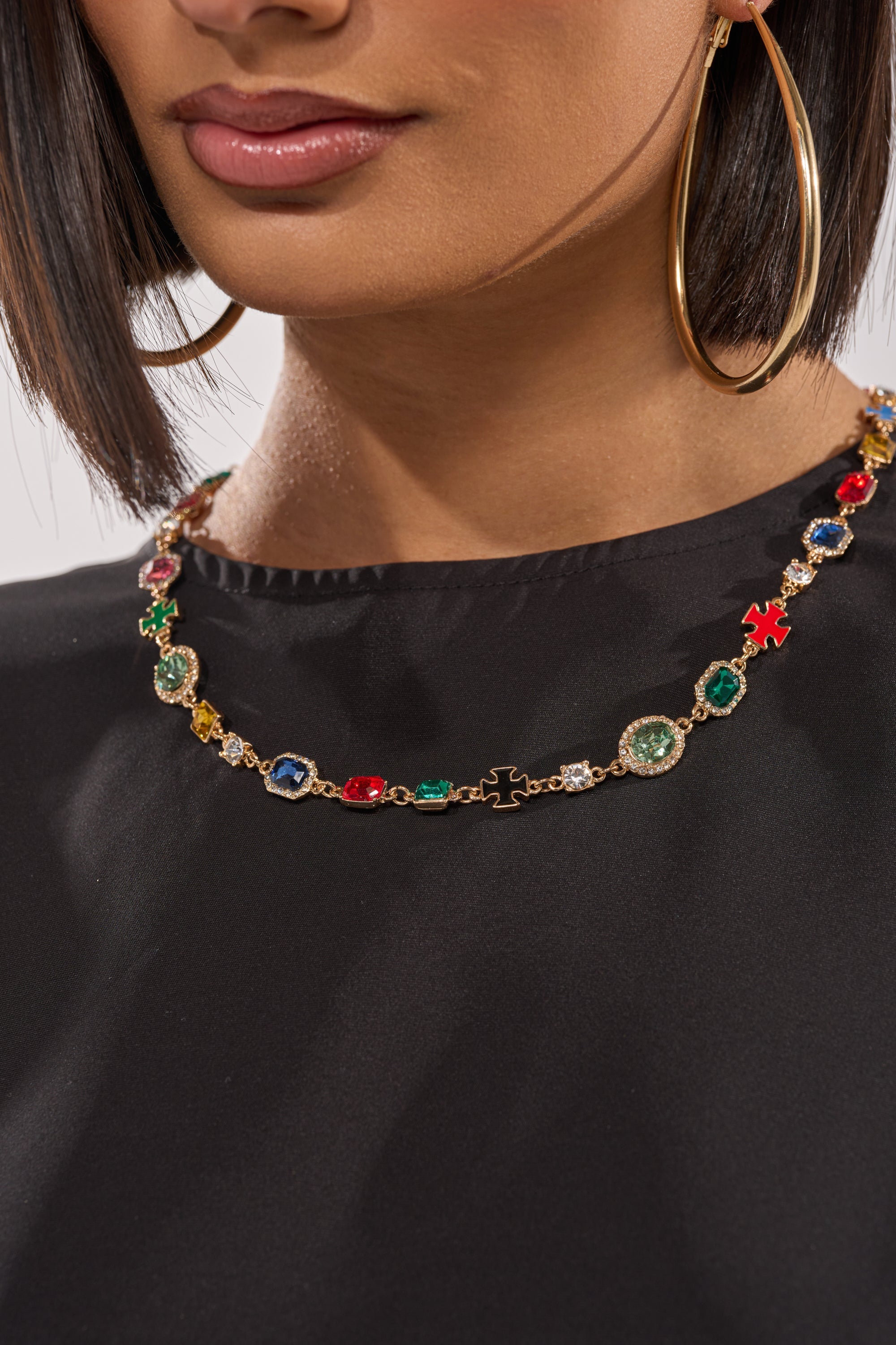 Wearing a black top and large gold hoops, a woman showcases the I AM HER NECKLACE—a colorful gemstone necklace with playful charms. Only her lips and upper chest are shown. 1