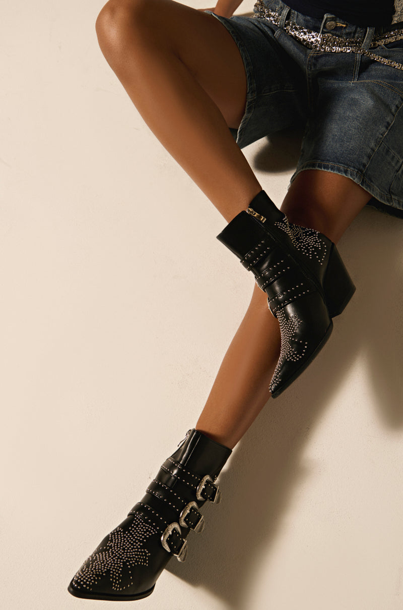 A person sits cross-legged, showcasing the AZALEA WANG CELENA BLACK WESTERN BOOTIE in studded vegan leather with three buckles. Paired with denim shorts against a plain background, it exudes modern flair and edgy charm.