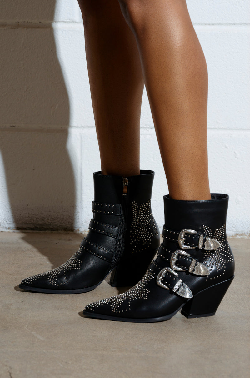 A person wears AZALEA WANG CELENA BLACK WESTERN BOOTIES, featuring black vegan leather, pointed toes, decorative silver studs, multiple metallic buckled straps, and block heels. The background has a white brick wall and a concrete floor.