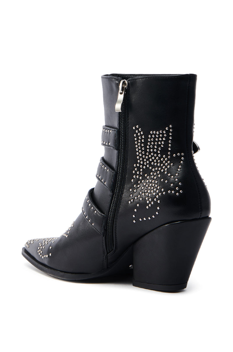 The AZALEA WANG CELENA BLACK WESTERN BOOTIE is a black ankle boot with a chunky heel crafted from vegan leather, featuring floral silver studs on the side, a visible zipper, and three decorative straps with metallic buckles across the front for a stylish western vibe.