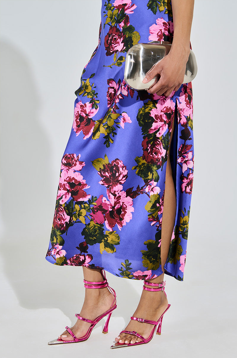 A person wears a vibrant floral dress featuring a thigh-high slit, paired with AZALEA WANG CELESIA FUCHSIA STILETTO SANDALS. They hold a silver metallic faux leather clutch. The dress bursts with purple, pink, and green hues on a blue backdrop.