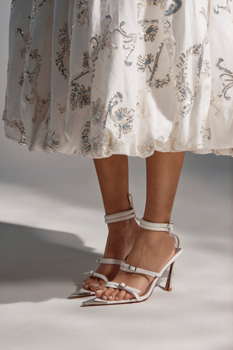 A woman stands in AZALEA WANG CELESIA WHITE STILETTO SANDALS, wearing an embellished white skirt. The image highlights her lower legs, the open toe silhouette of the sandals, and the detailed skirt hem.
