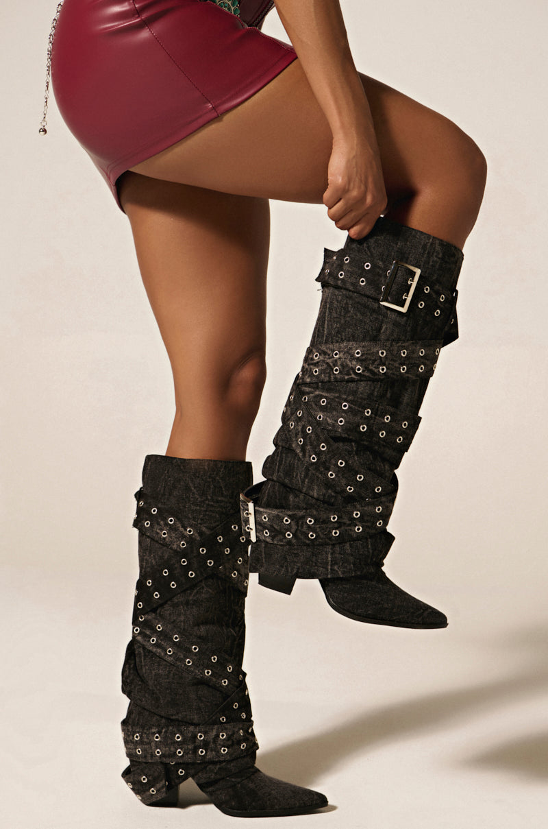 A person in a short, shiny burgundy skirt and knee-high CENSORED BLACK DENIM BELTED CHUNKY BOOT with silver buckles and grommets balances on one foot to adjust the other boot against a plain, light-colored background.