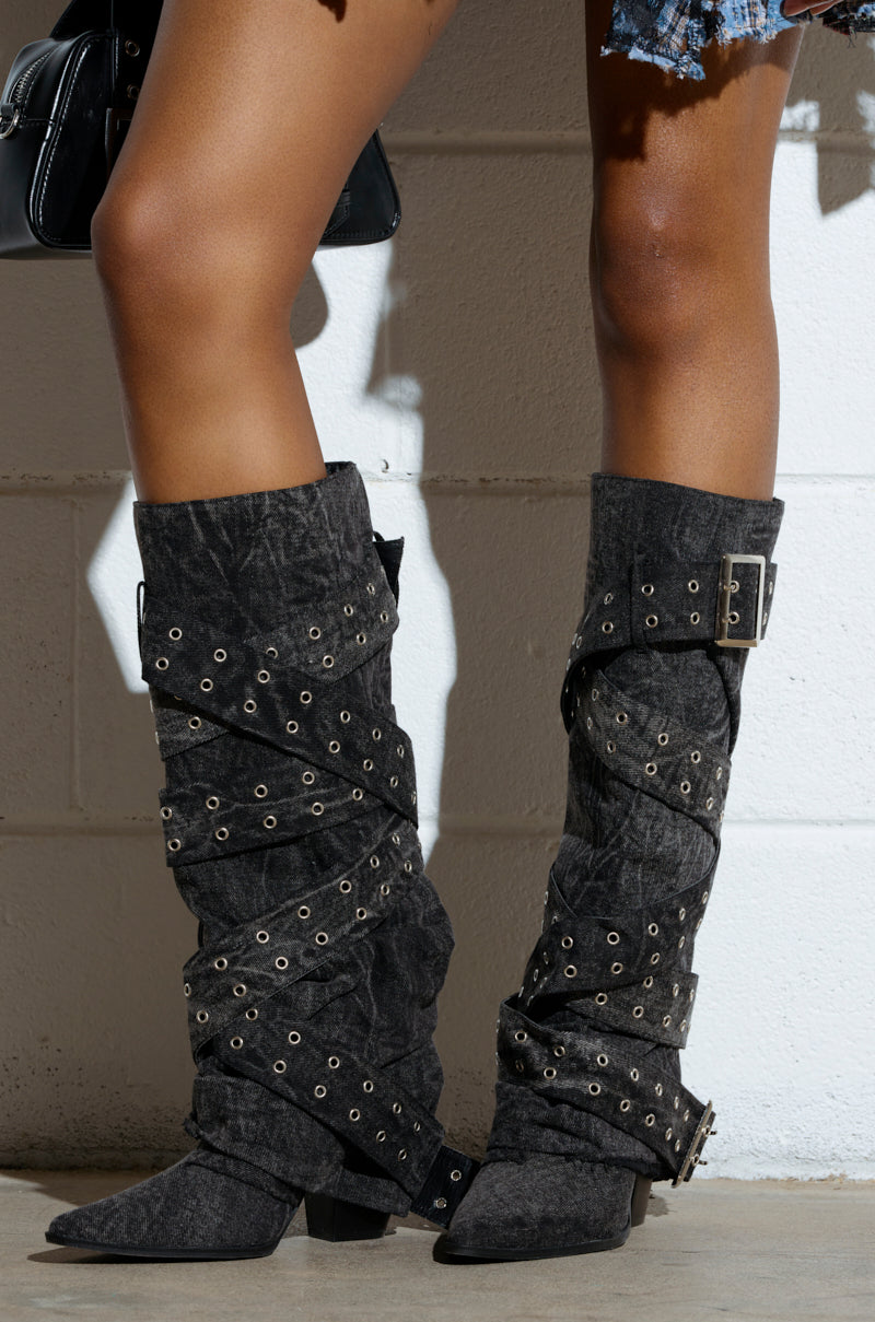A person showcases the CENSORED BLACK DENIM BELTED CHUNKY BOOT, featuring black, slouchy knee-high design with grommets, buckles, pointed toes, and low chunky heels. They're paired with denim shorts and a black handbag against a white brick wall backdrop.
