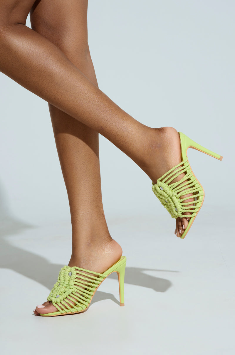The AZALEA WANG CERES LIME ROPE WITH RHINESTONE BEADS SANDAL, featuring intricate braided straps and stiletto heels, is worn on feet with crossed ankles against a light blue background.