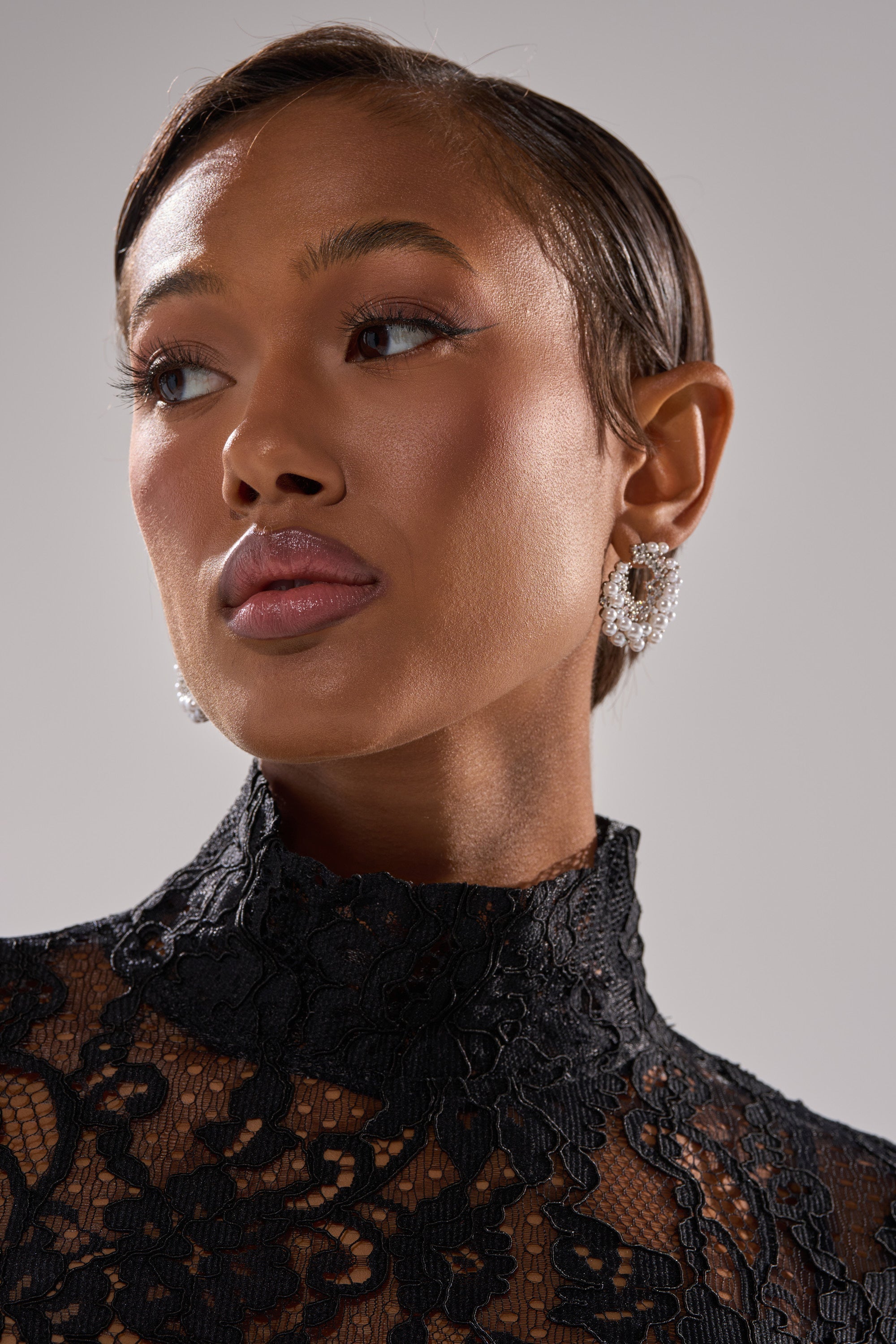 Woman with short, slicked-back hair wears the TRISH EARRING and a high-neck black lace top, looking slightly to the side against a neutral background. Her makeup is natural with glossy lips and defined brows. 1