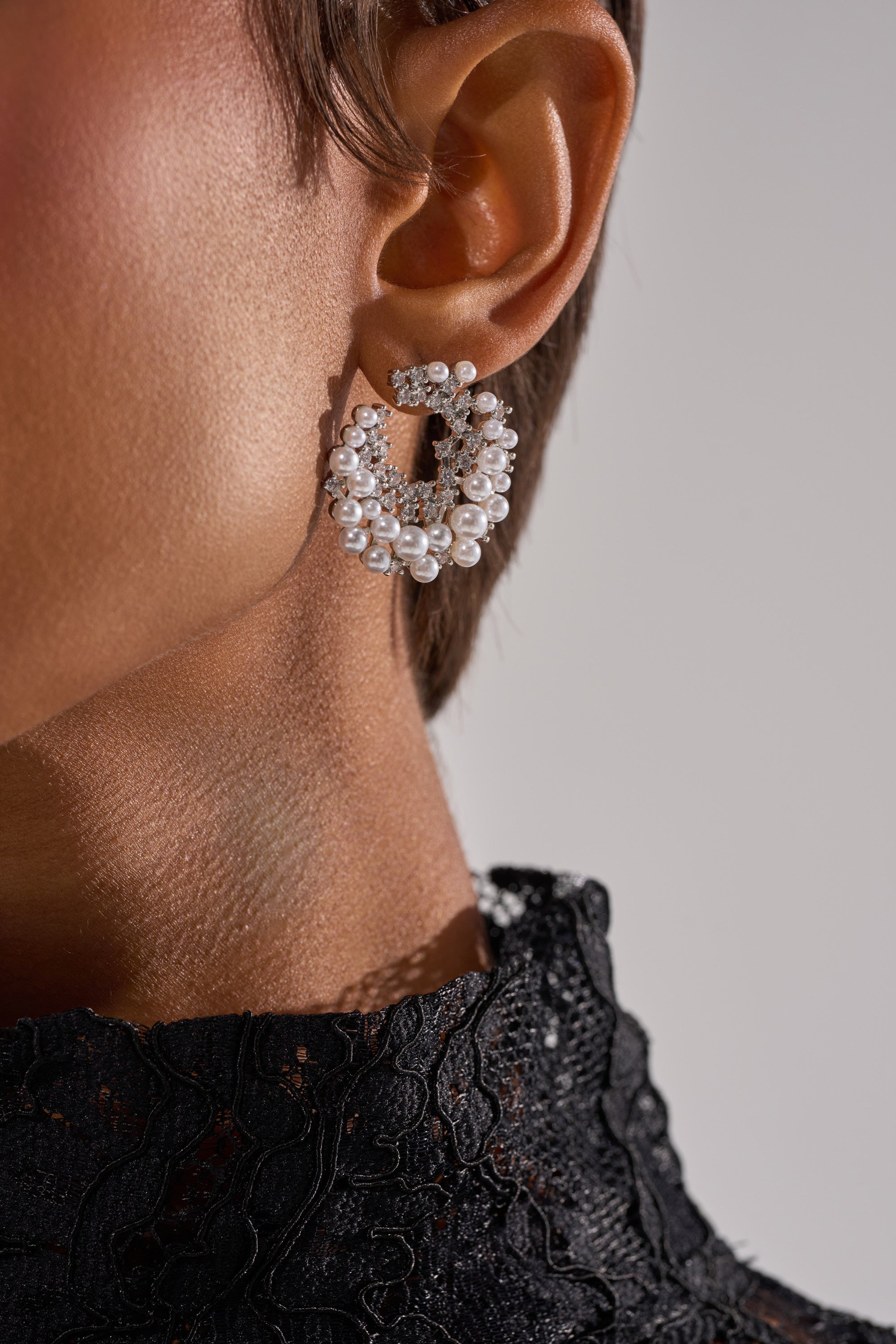 Close-up of a person with short hair in a black lace outfit, wearing the TRISH EARRING—a large circular statement piece with rhinestones, pearls, and crystals. The neutral out-of-focus background highlights this bold accessory. 3