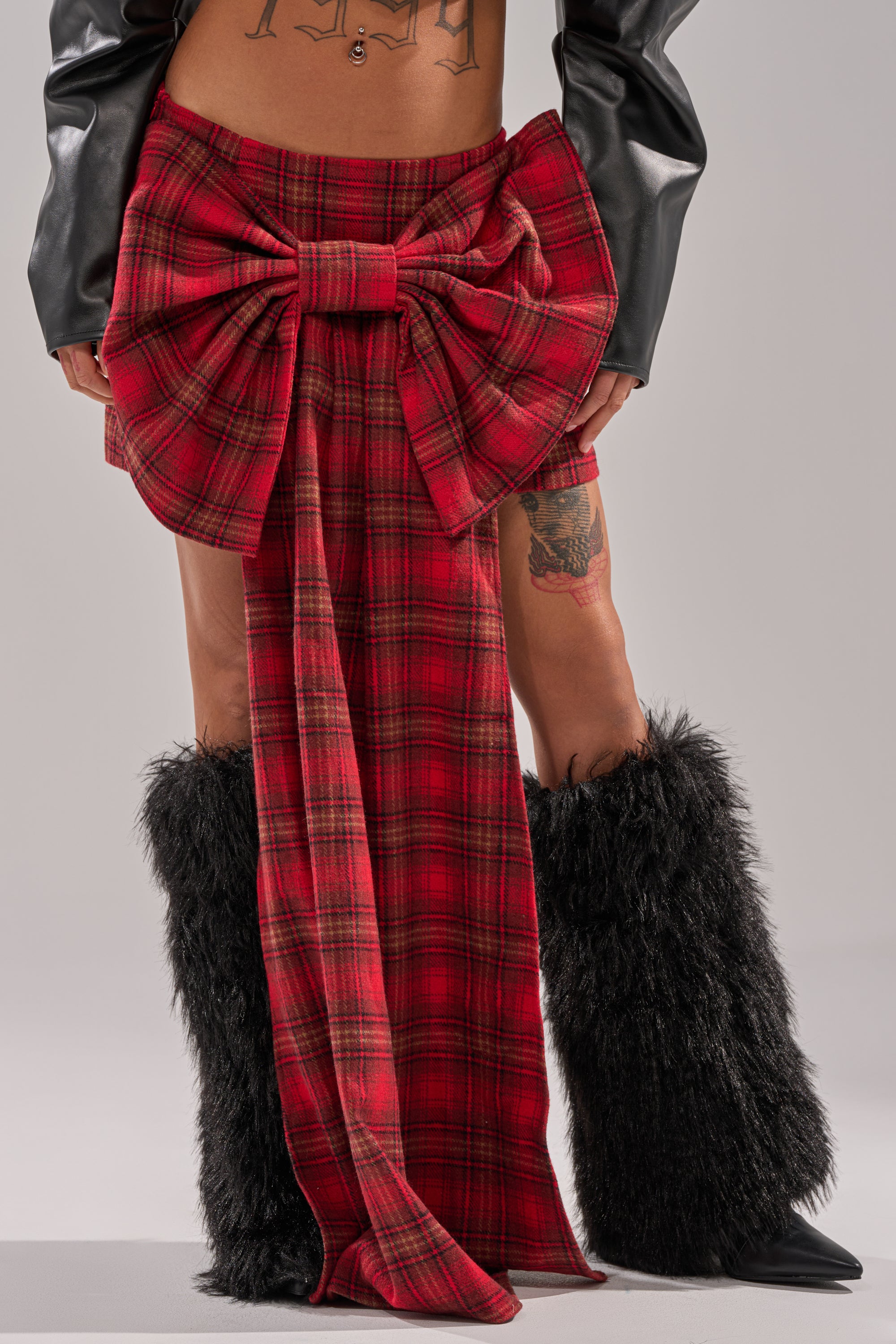 Wearing the LOVELY DAYS PLAID BOW MINI SKIRT with a large front bow, black faux fur leg warmers, and a black leather jacket, the person's midriff tattoo is visible in this cropped shoulders-to-feet image. 1