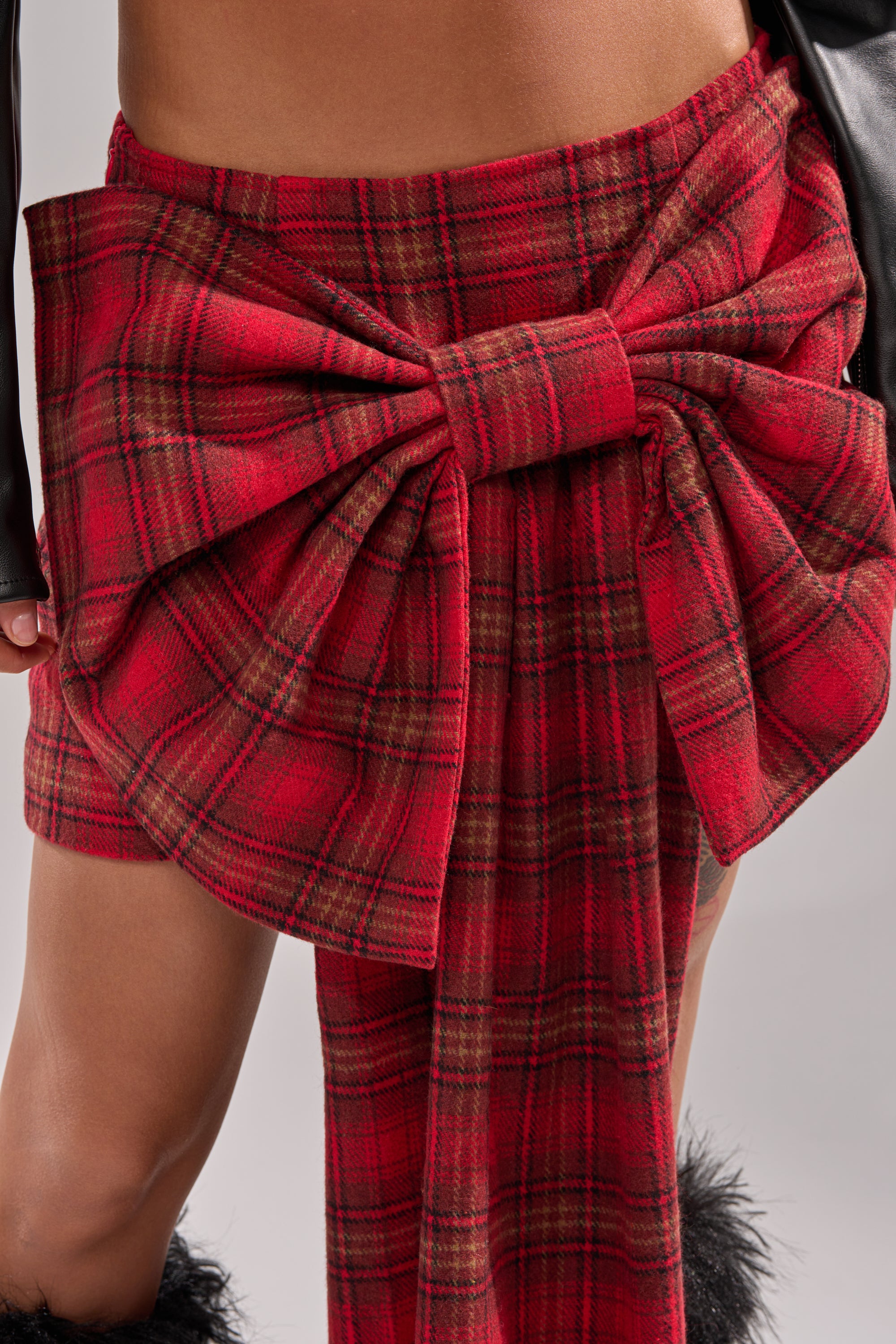 A close-up of a person wearing the LOVELY DAYS PLAID BOW MINI SKIRT, featuring a bold front bow. Their bare midriff and leg are shown, with hints of a black leather sleeve and black furry shoes also visible. 8