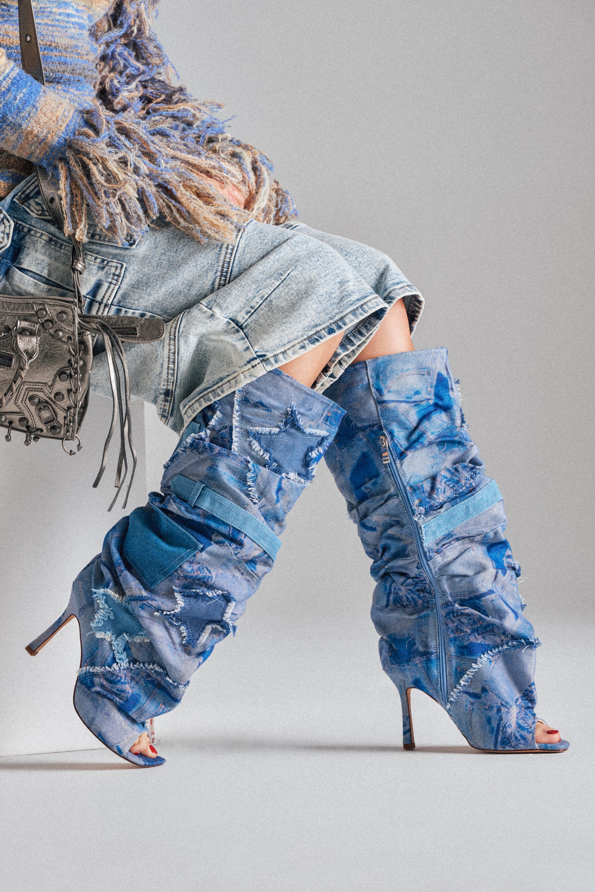 A person wearing a frayed denim skirt, a blue and beige fringed sweater, and the AZALEA WANG CHAITRA DENIM OPEN TOE BOOT sits cross-legged, with a silver metallic handbag at their side. 1