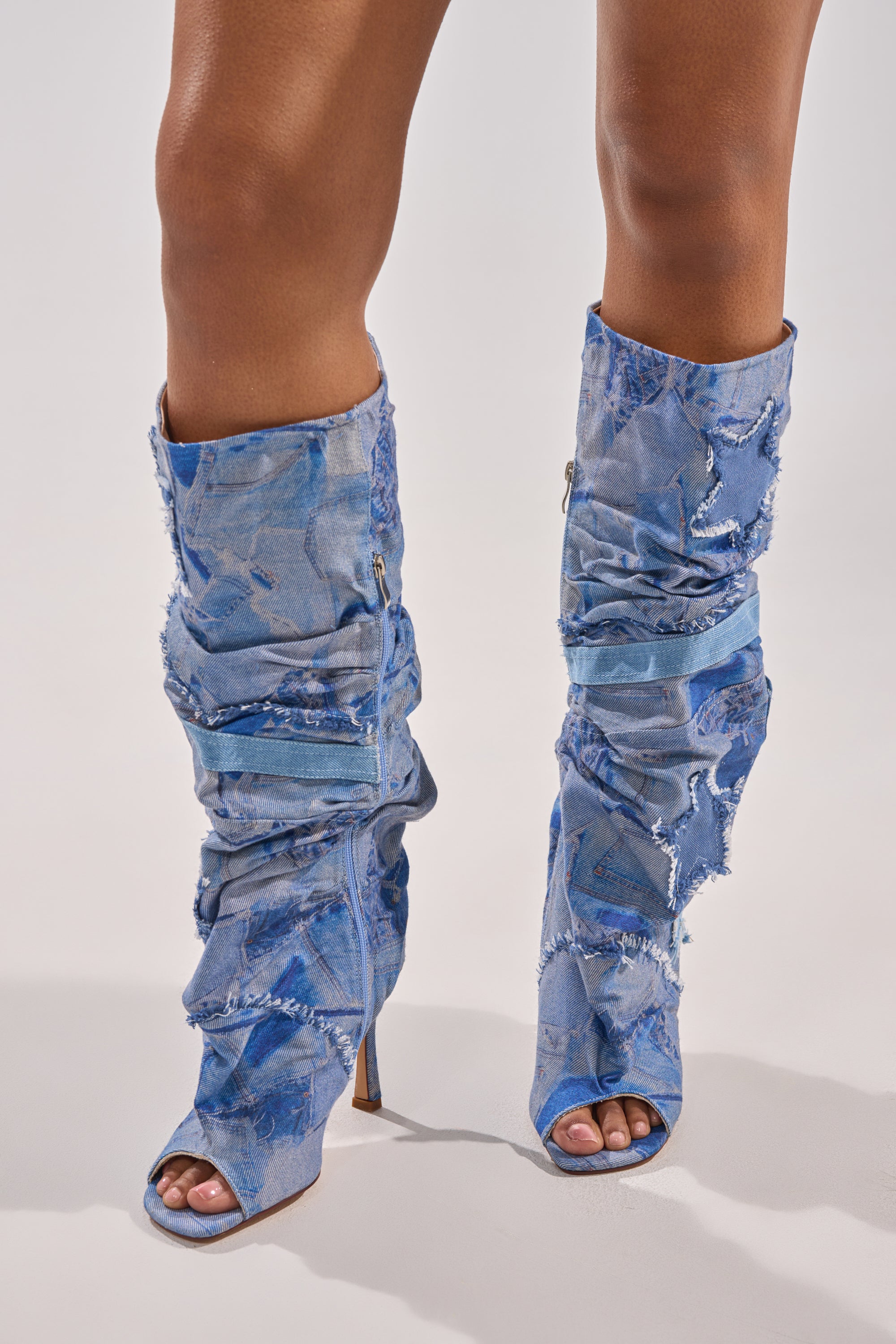A person wearing the AZALEA WANG CHAITRA DENIM OPEN TOE BOOT—knee-high slouch boots made from distressed blue denim with frayed edges—stands on a light surface.