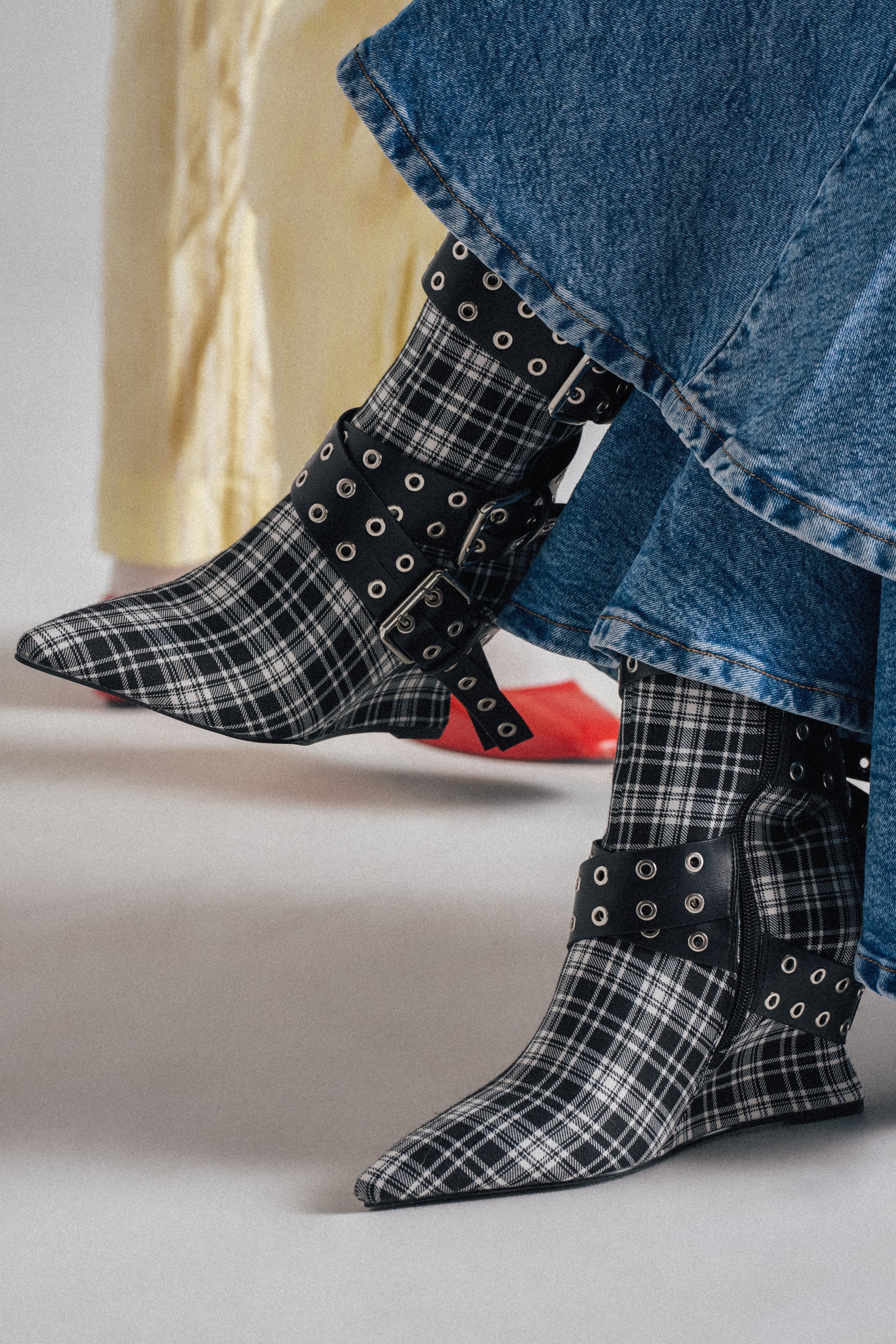 Two people stand together; one wears red shoes and yellow pants, while the other sports a blue denim skirt with AZALEA WANG CHALKIS BLACK WHITE PLAID BUCKLE WEDGE BOOTIE featuring grommet straps and pointed toes. 0