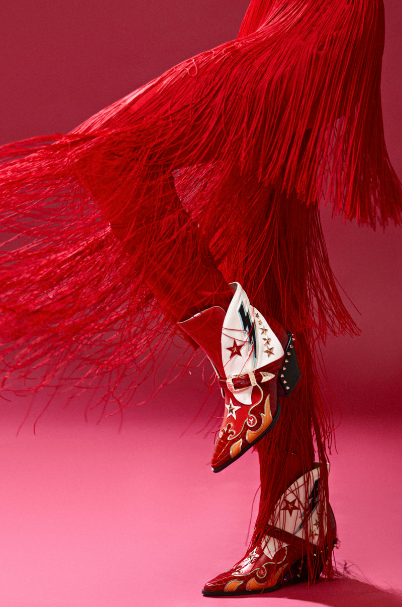 An individual is sporting AZALEA WANG CHANNING MULTI WESTERN BOOTIE, flaunting striking faux leather cowboy boots with star and flame motifs. These bold booties stand out against solid red fringe pants and feature an eye-catching chunky heel.