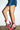 A person in a short denim skirt and the AZALEA WANG CHANNY MULTI BOOTIE GRAPHIC WESTERN WITH STUDS with chunky heels stands before a white brick wall.