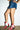 A person stands against a white brick wall wearing a short blue denim skirt and the AZALEA WANG CHANNY MULTI BOOTIE GRAPHIC WESTERN WITH STUDS, featuring star and moon designs, with hands on hips and bright red nails showing.
