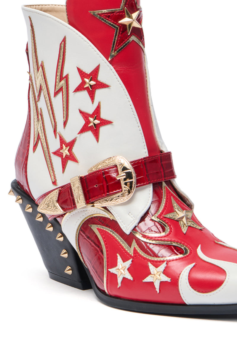 A close-up of the AZALEA WANG CHANNY MULTI BOOTIE GRAPHIC WESTERN WITH STUDS shows its red, white, and gold faux leather design with stars, lightning bolts, a gold buckle, gold spikes, and a chunky black heel.