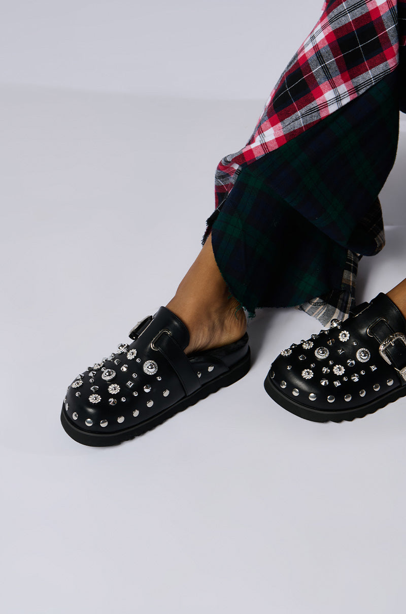 A person wearing AZALEA WANG CHATTER FUR LINED MULEs, adorned with silver studs and gems. They are dressed in plaid-patterned pants featuring shades of red, black, green, and white against a plain and light-colored background.