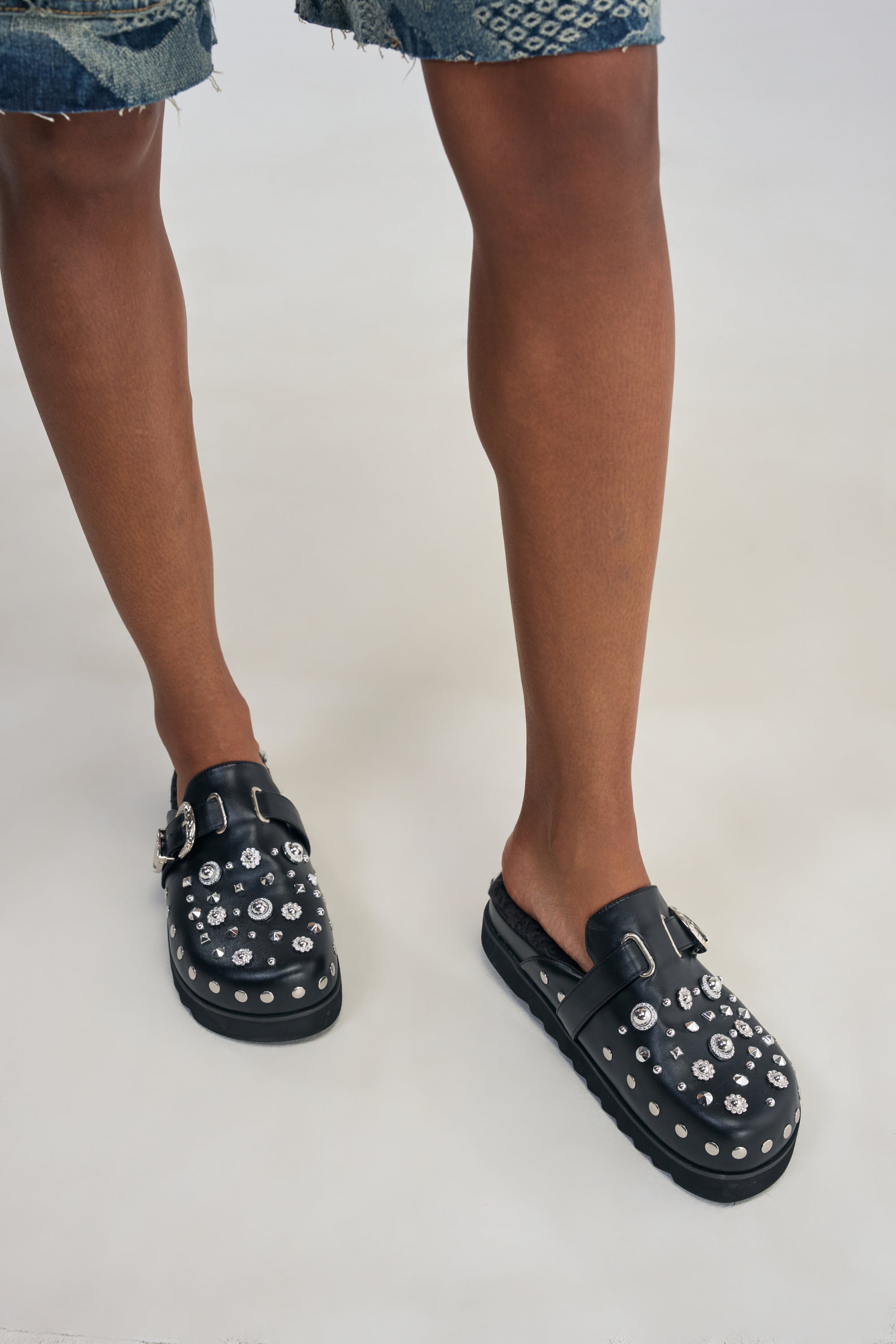 Wearing the AZALEA WANG CHATTER FUR LINED MULE—studded black slip-on shoes with buckles reminiscent of black faux leather clogs—a person stands on a light background, paired with frayed, patchwork denim shorts.