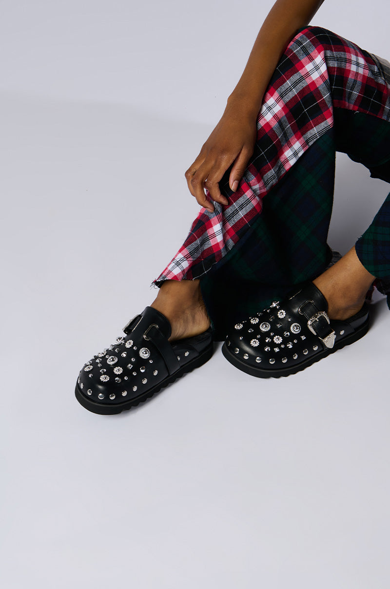 A person wearing the AZALEA WANG CHATTER FUR LINED MULE, characterized by its metallic studs and buckles, is seated with one leg slightly bent. They are also dressed in plaid pants featuring a red, white, and black pattern layered over a green, blue, and black one for added style. These mules have a comfort cushion insole for enhanced comfort.