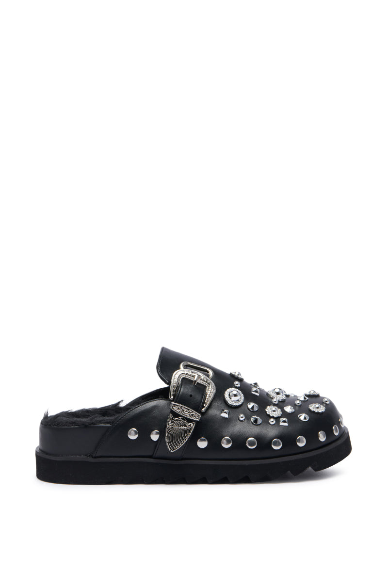 The AZALEA WANG CHATTER FUR LINED MULE is a black faux leather mule featuring silver studs, rhinestones, and various decorative embellishments. This slip-on shoe showcases a prominent buckle on the upper side and boasts a ridged, chunky rubber sole for added style and stability, complemented by a comfort cushion insole to enhance each step.