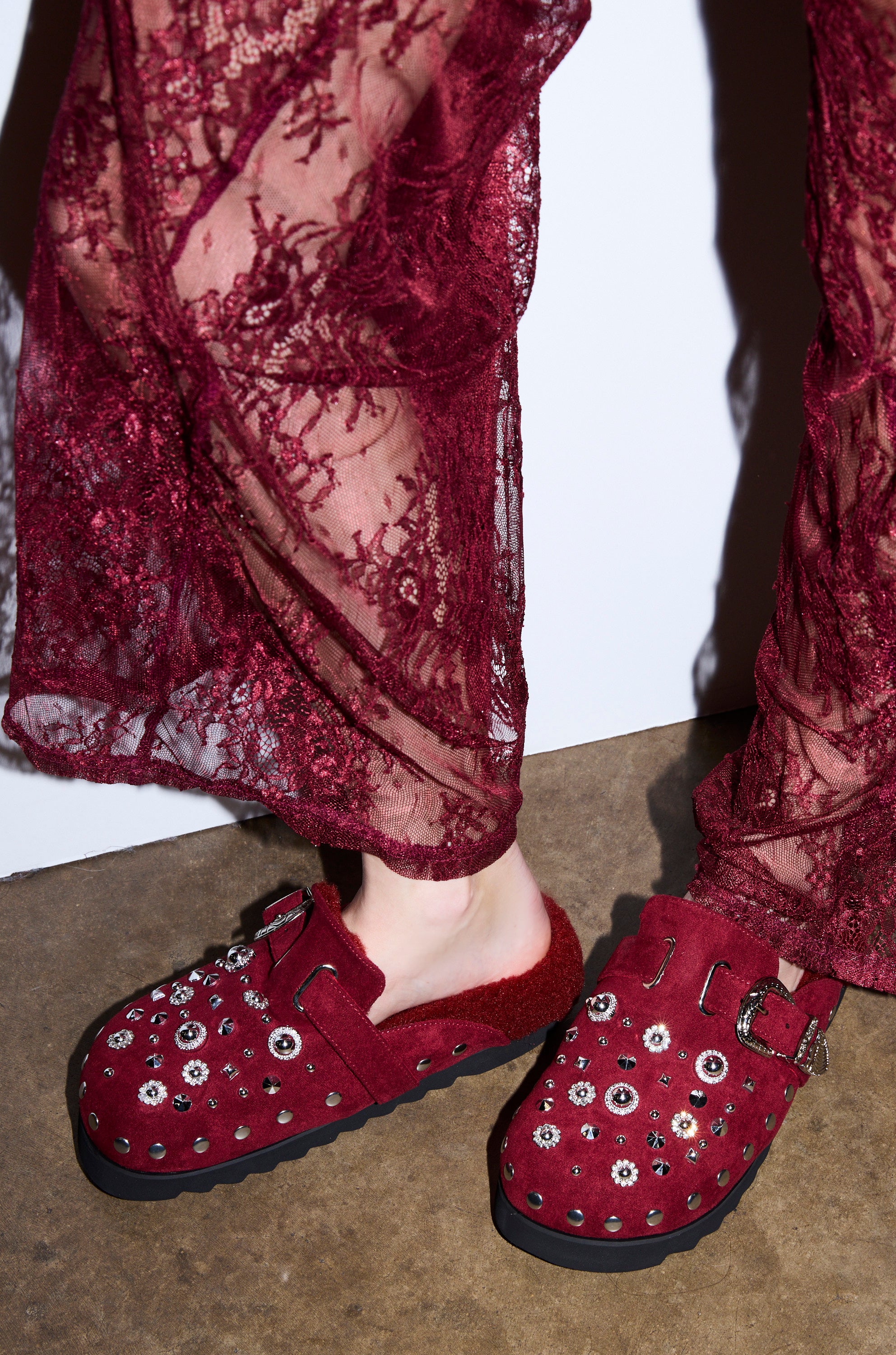 A person wearing sheer burgundy lace pants and AZALEA WANG CHATTER BURGUNDY FUR SLIP ON CLOGS stands on a brown floor against a white wall.