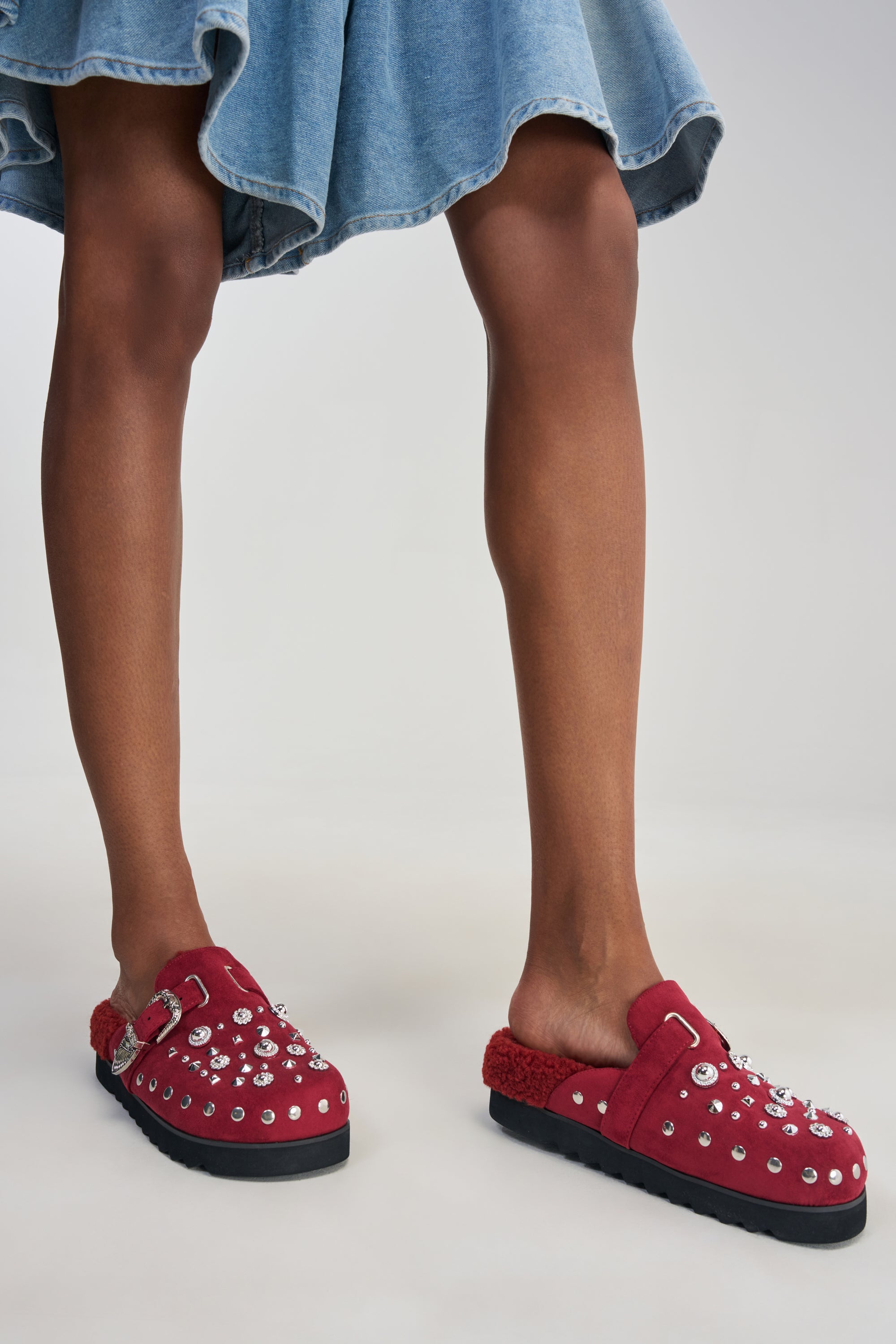 Wearing a blue denim skirt and red slip-on shoes adorned with metallic studs and buckles, the individual is complemented by the AZALEA WANG CHATTER BURGUNDY FUR SLIP ON CLOG, which features cozy sherpa lining, as they stand on a light background.