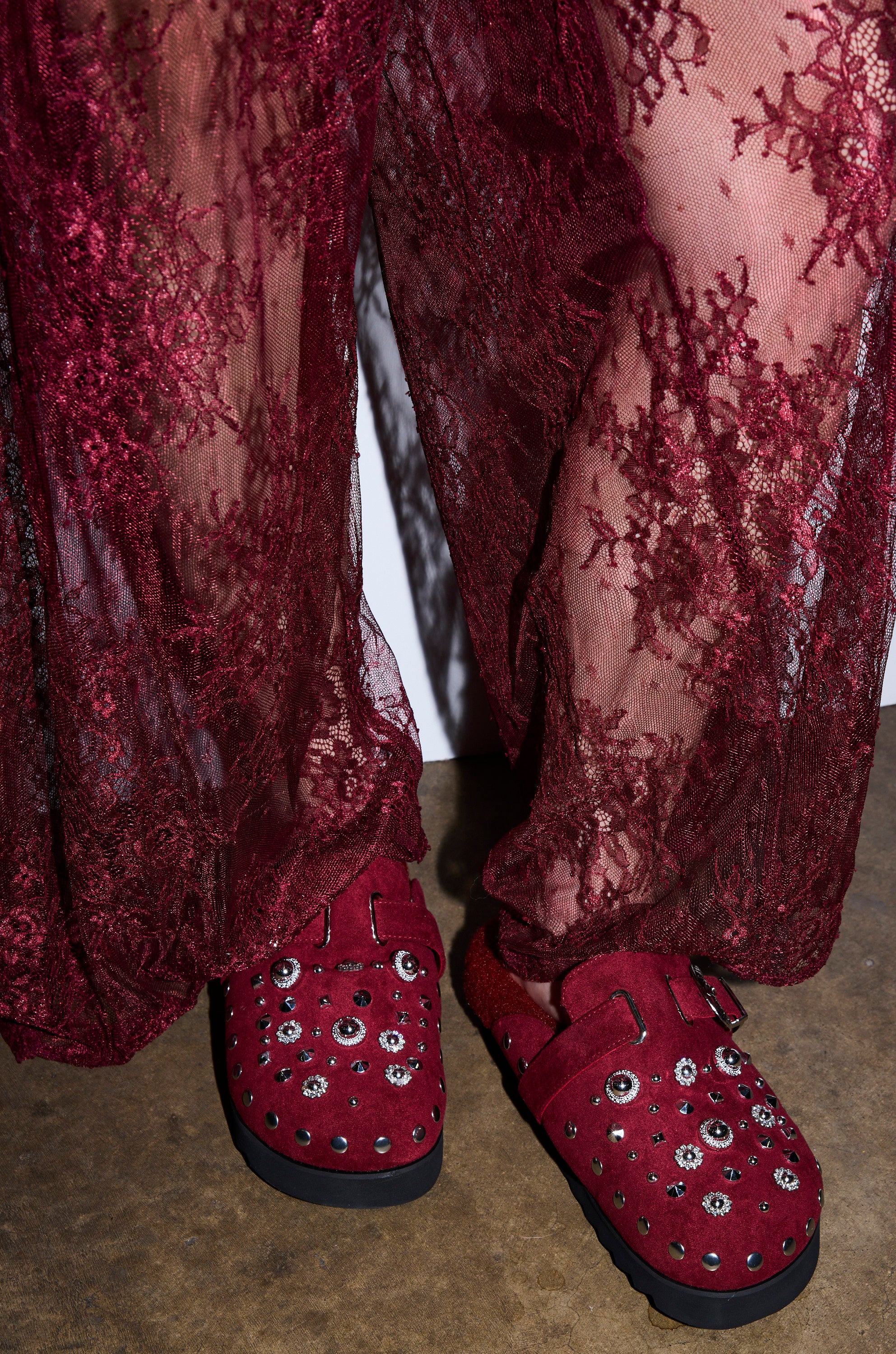 Close-up of a person wearing sheer, burgundy lace pants and AZALEA WANG CHATTER BURGUNDY FUR SLIP ON CLOGS with faux suede and silver studs, standing on a textured brown floor.
