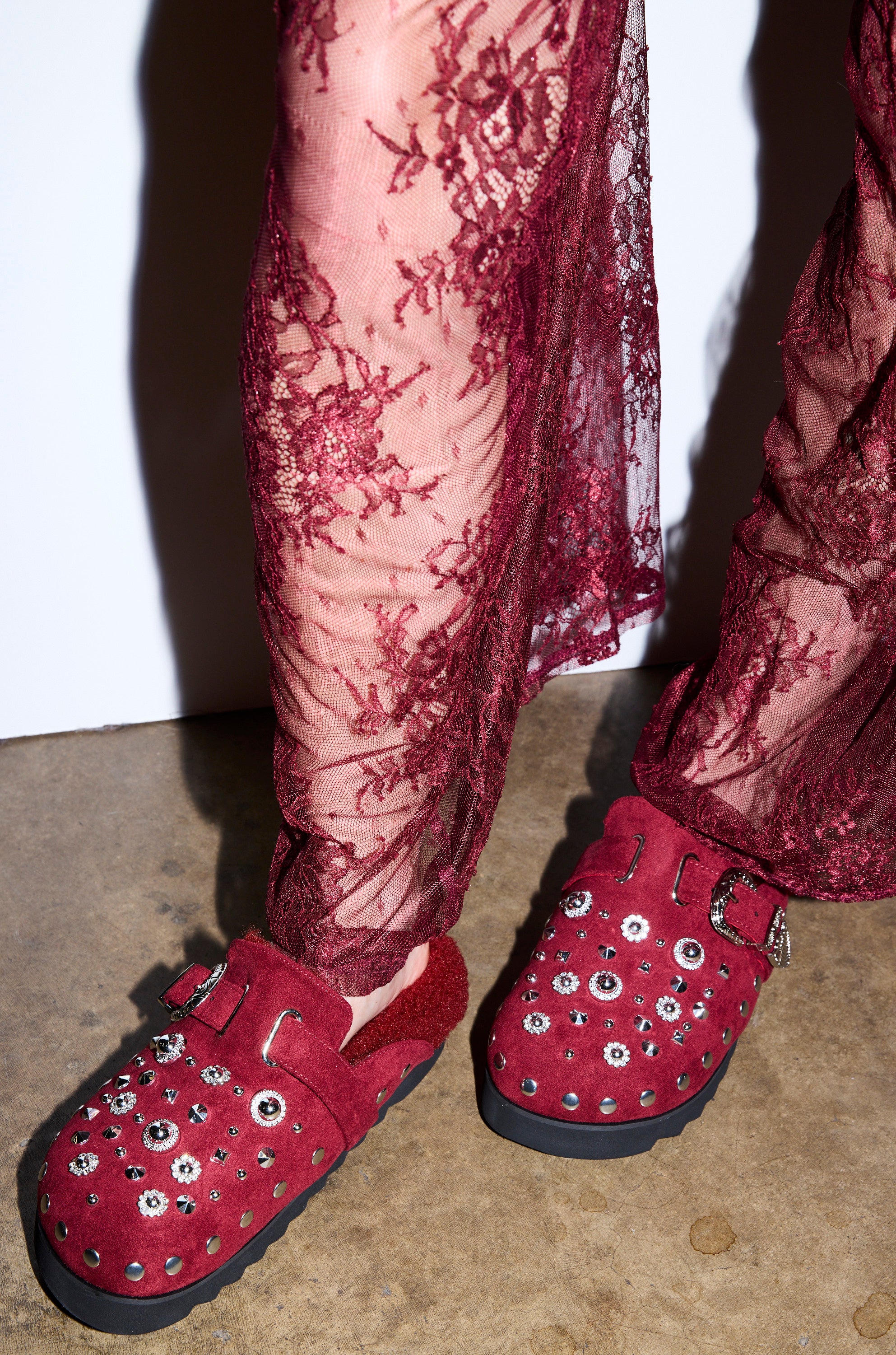 A person wearing sheer burgundy lace pants and AZALEA WANG CHATTER BURGUNDY FUR SLIP ON CLOGS with silver studs stands on a concrete floor.