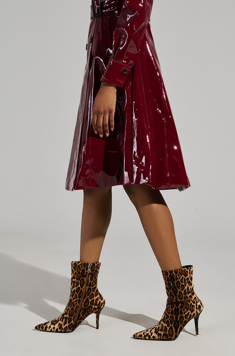 A person wearing a shiny burgundy trench coat and the AZALEA WANG CHAUNDRA Leopard Embellished Stiletto Bootie stands against a plain background. The outfit is stylish, with the boots adding a bold touch. 0